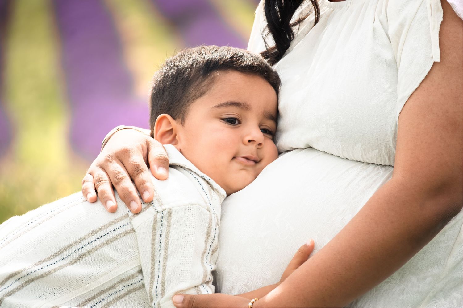 A young boy lovingly rests his head on his pregnant mother's belly outdoors in a lavender field.