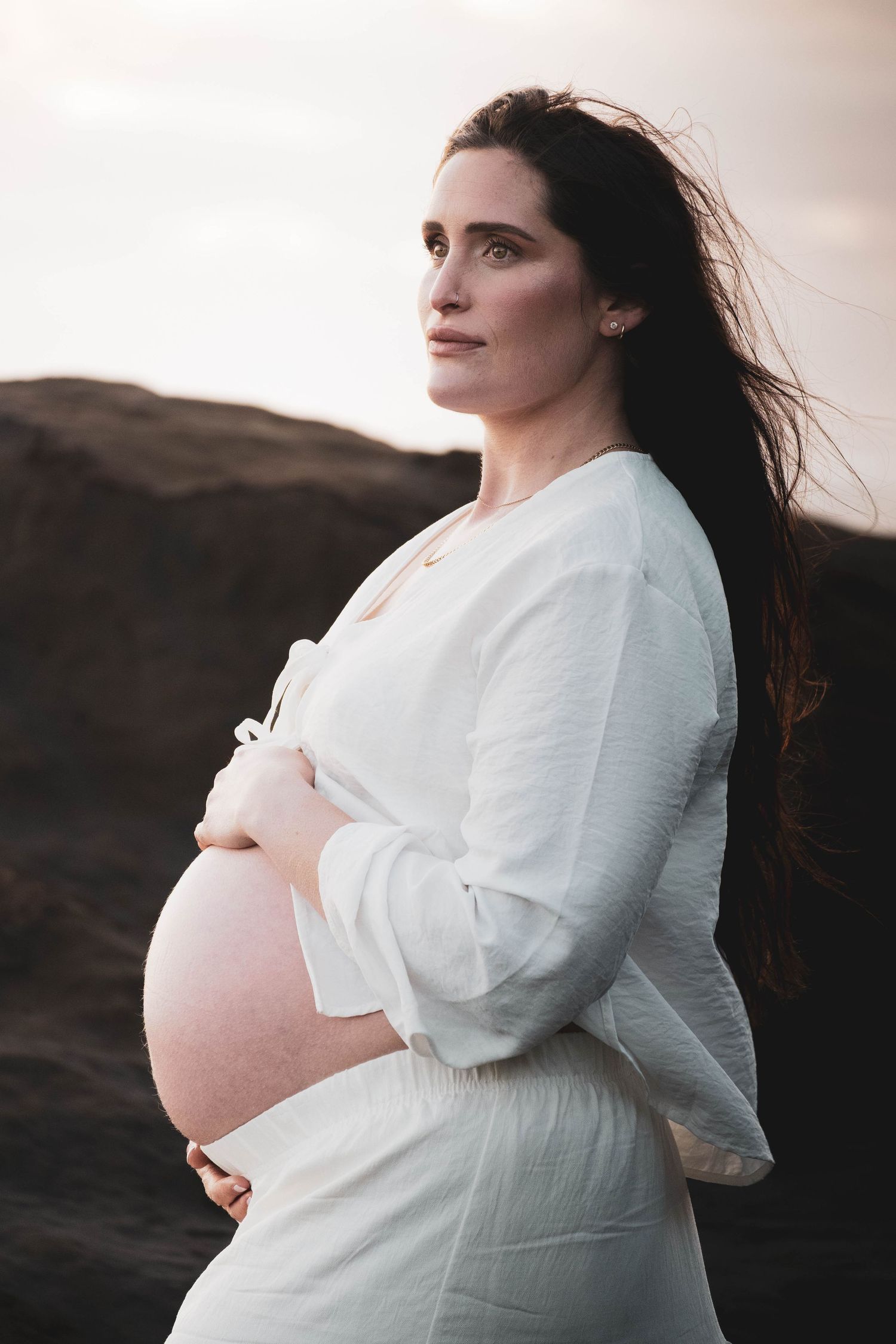 Pregnant woman in white dress cradling baby bump outdoors at sunset with dark rocky background.