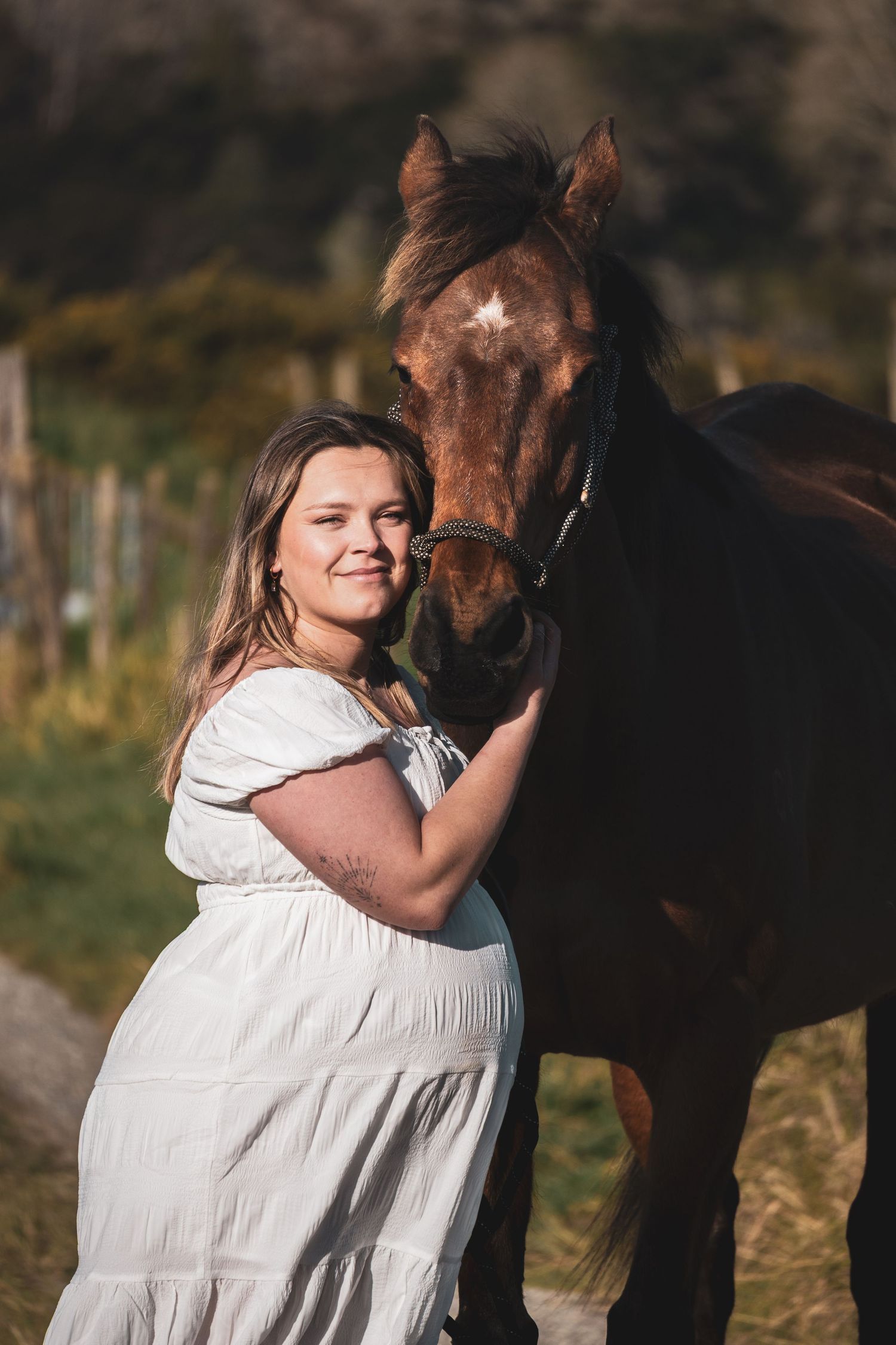 Woman in white dress smiling beside a brown horse in a sunny outdoor field setting.