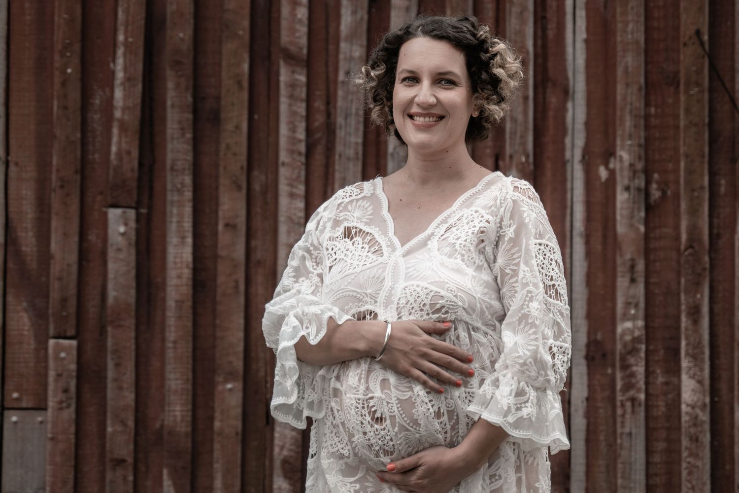 Smiling pregnant woman in white lace maternity dress cradling baby bump outdoors against rustic wooden wall.