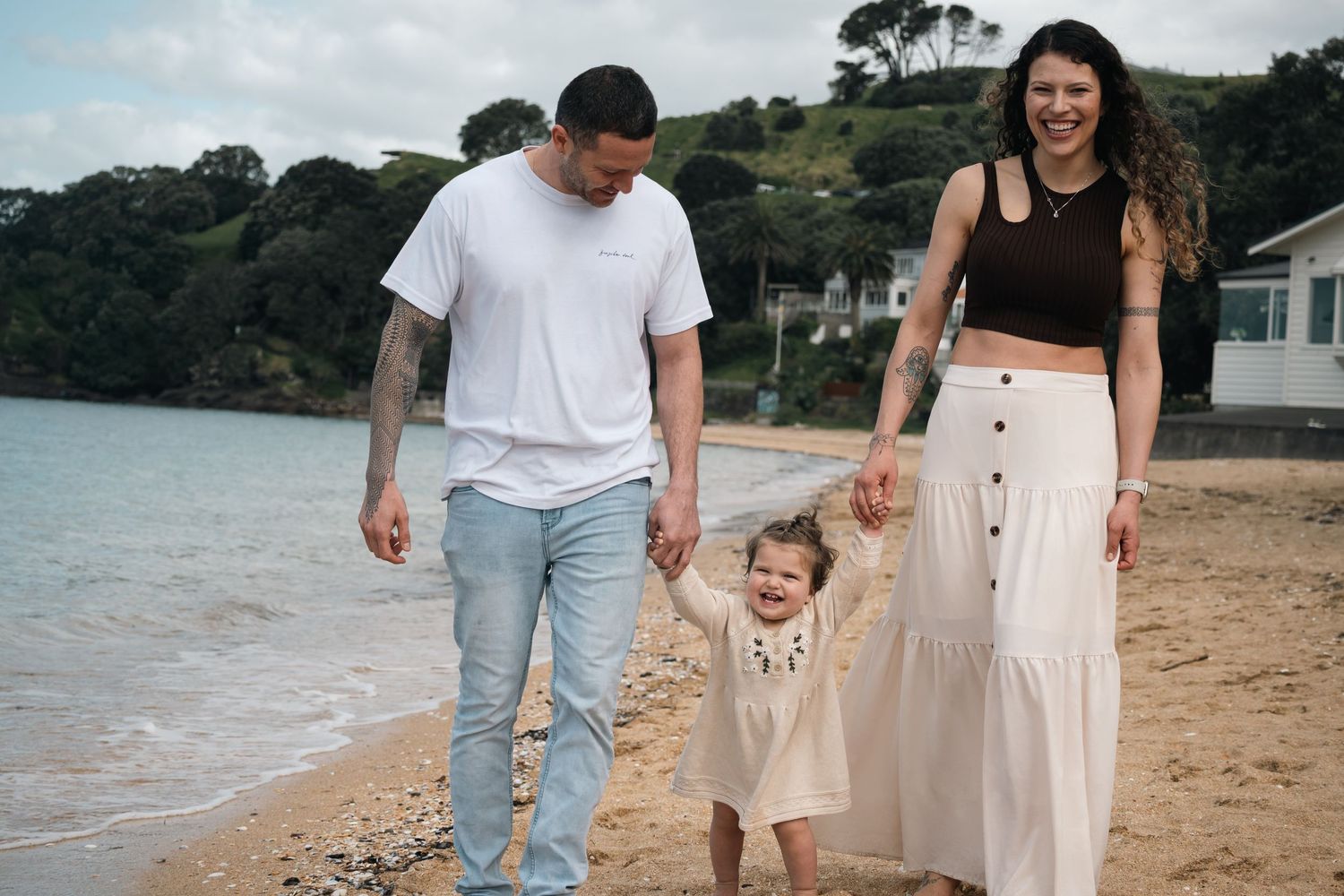 A happy family of three walks hand-in-hand on a sandy beach, parents swinging their toddler between them.