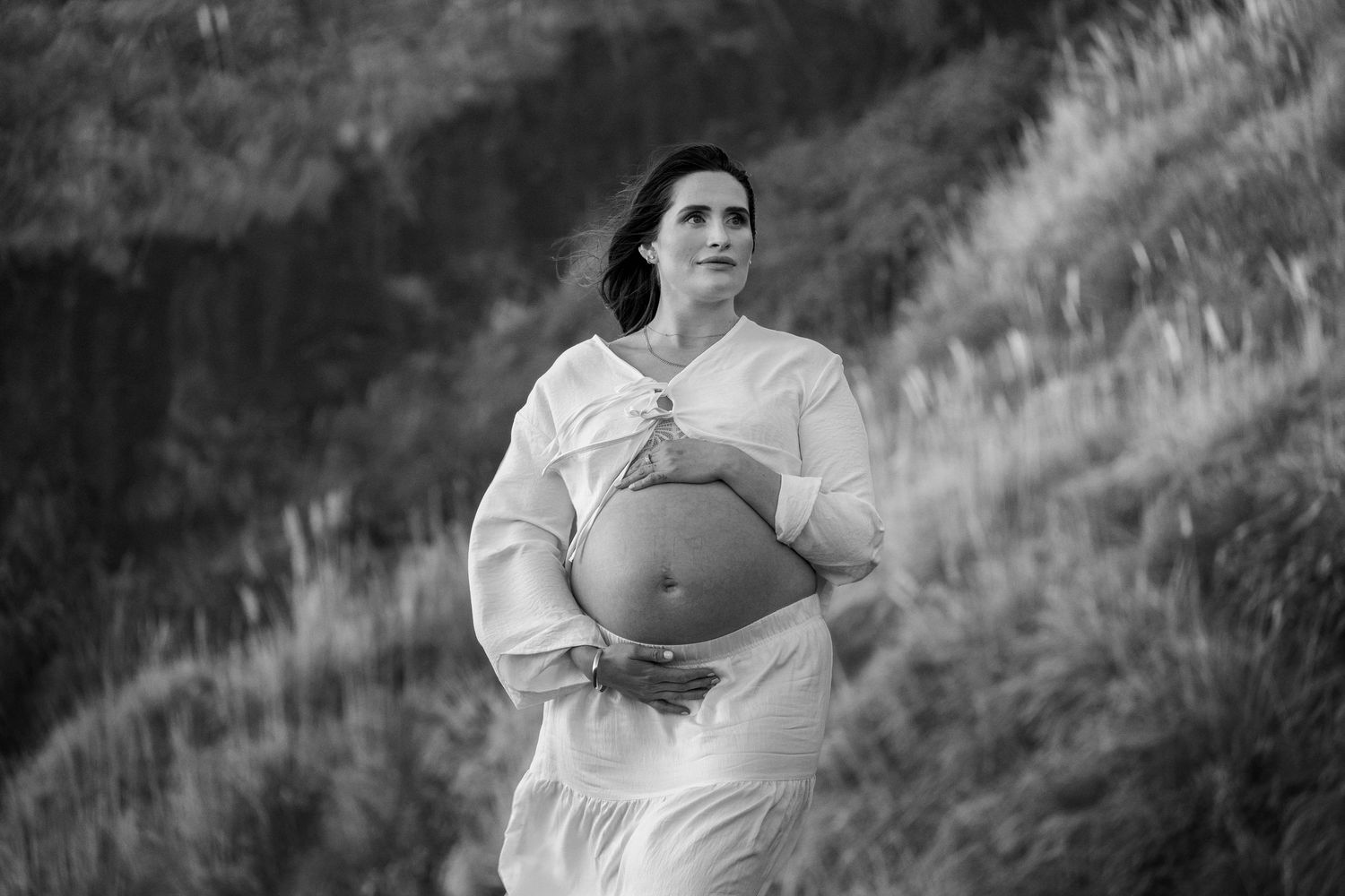 Black and white maternity photo of a pregnant woman in a white dress holding her belly outdoors.