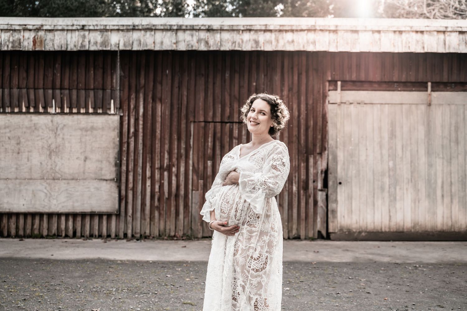 Pregnant woman in white lace gown holding baby bump, standing outdoors near rustic barn wall in soft sunlight.