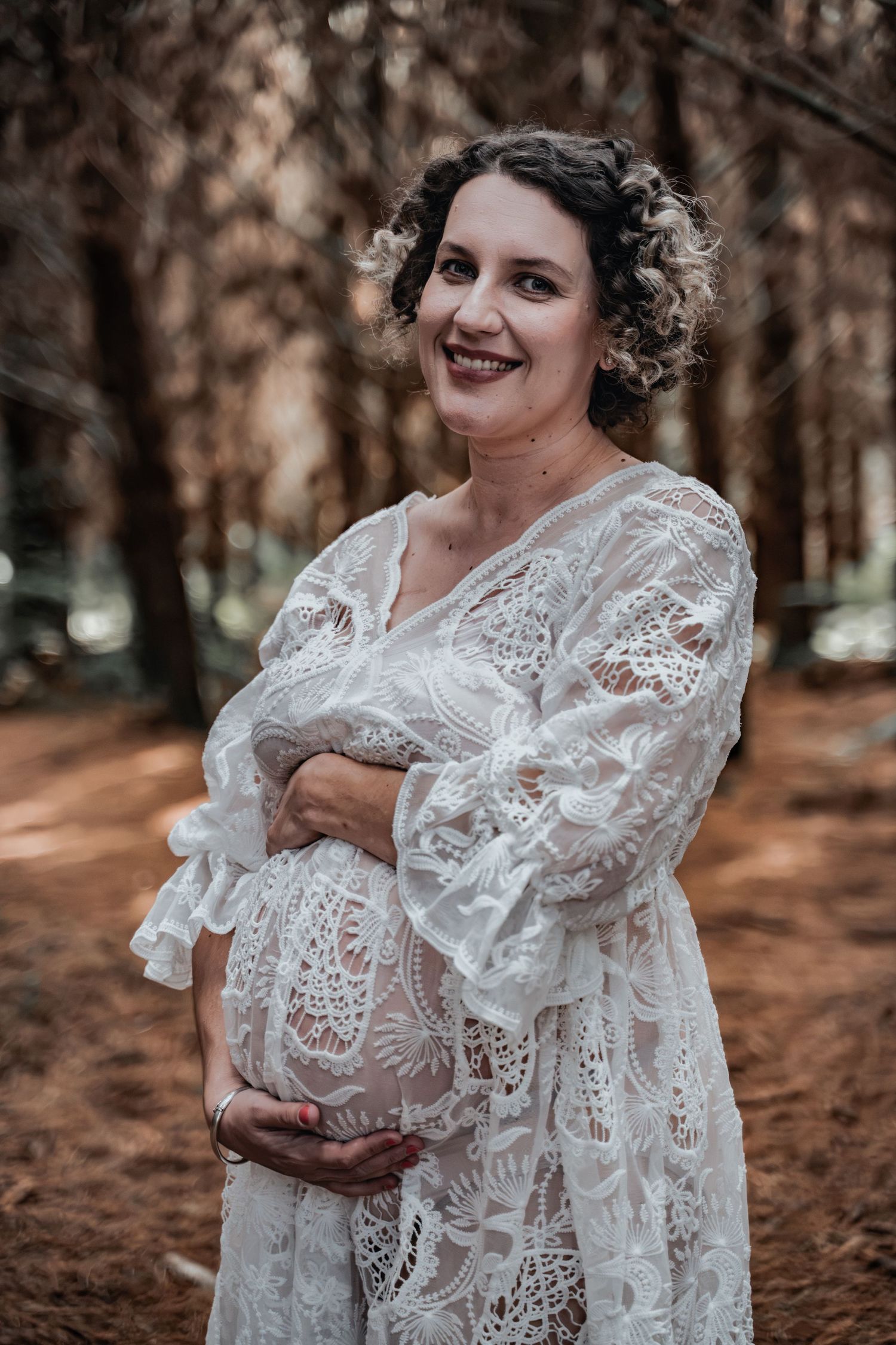 Smiling pregnant woman in white lace dress cradling her baby bump in a forest setting.
