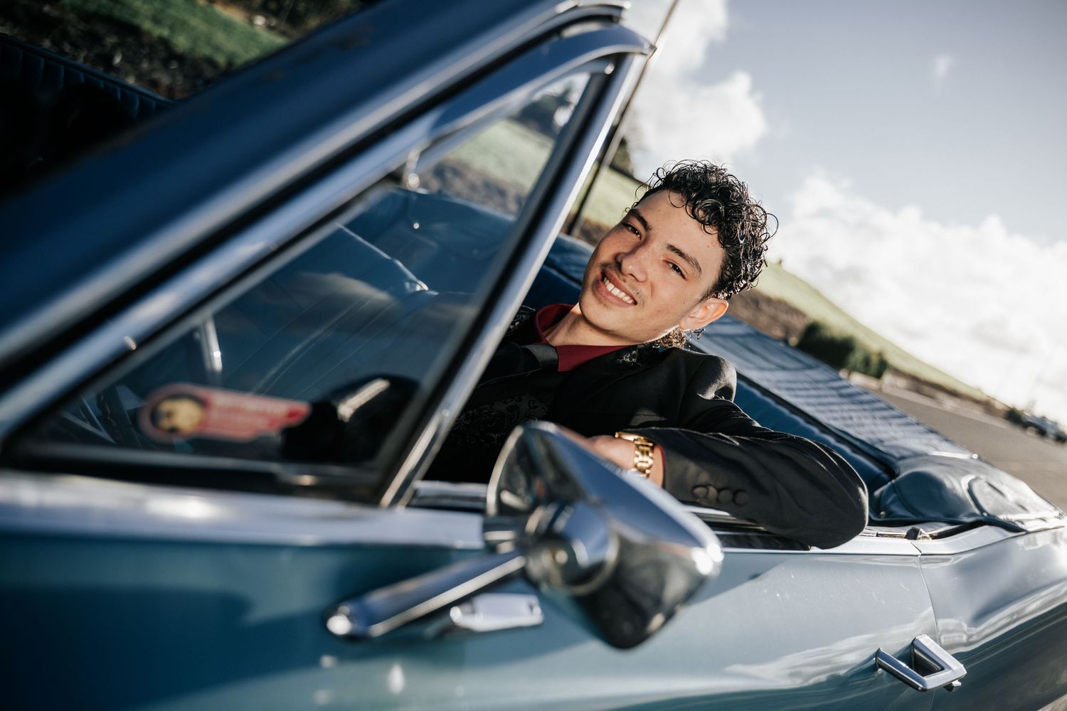 Young man with curly hair smiling while leaning out of a classic blue convertible car on a sunny day.