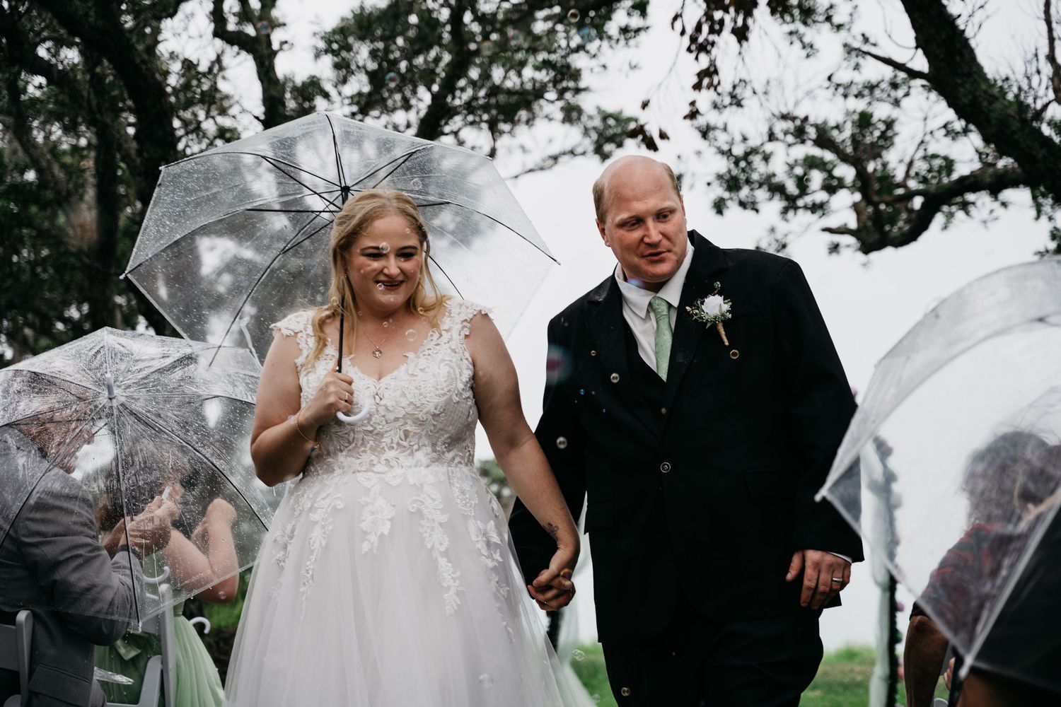 Bride and groom walk under clear umbrellas during a romantic outdoor rainy auckland wedding ceremony.