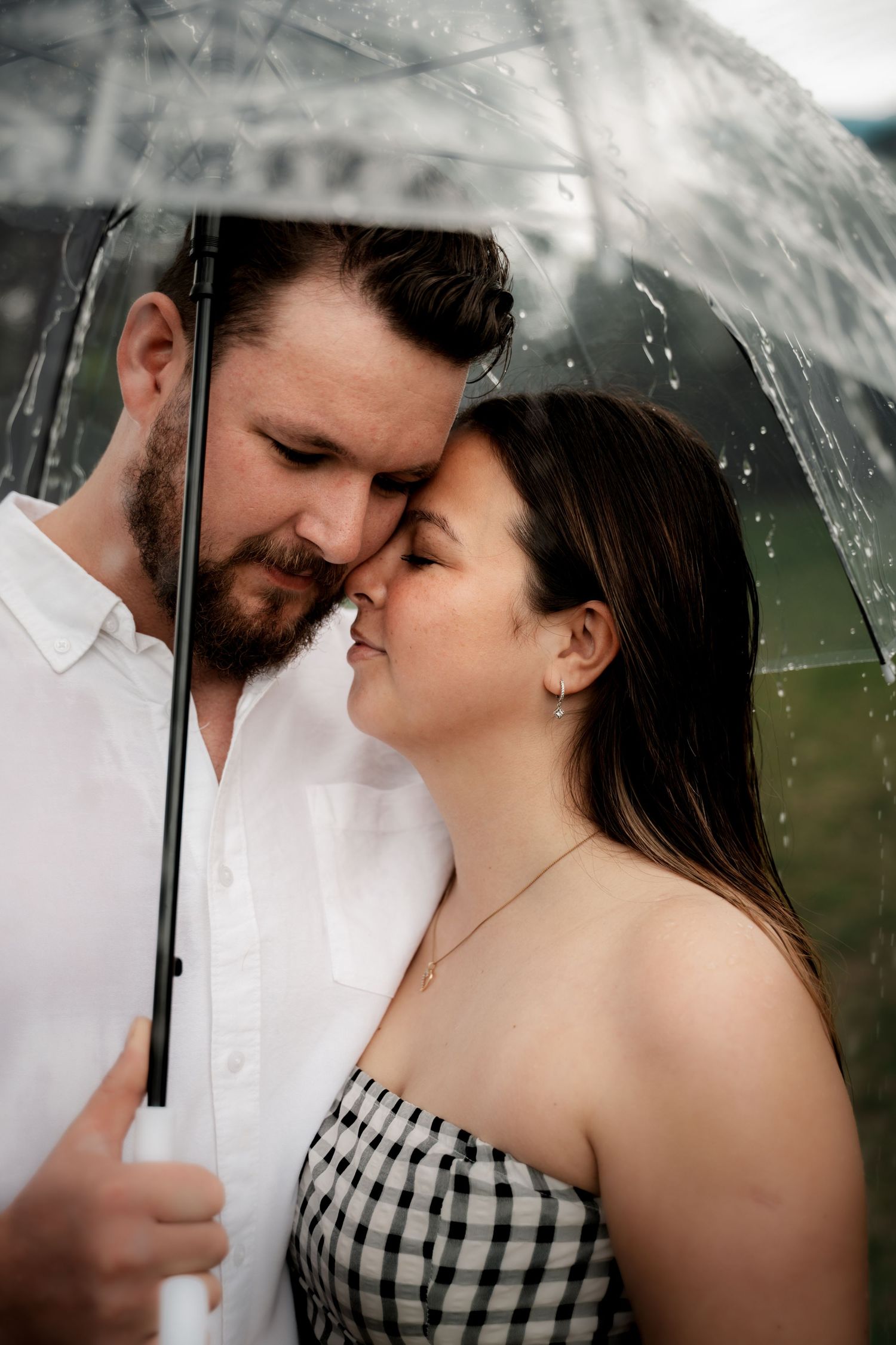 A couple shares an intimate moment under a clear umbrella in the rain, the man in a white shirt nuzzling the woman.