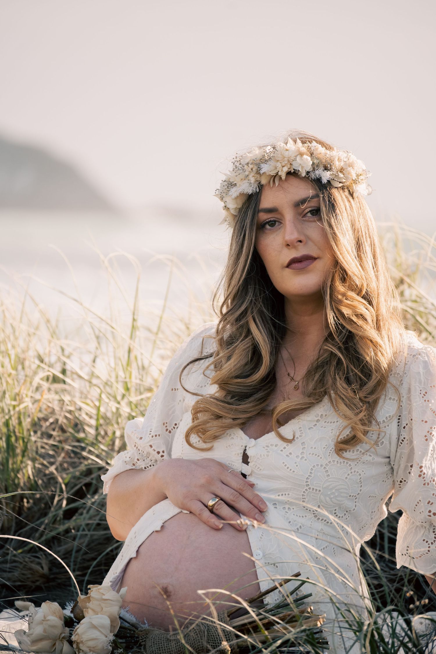 Pregnant woman in white lace dress and floral crown sits among beach grass at sunset on Muriwai Beach for her Maternity Photoshoot.