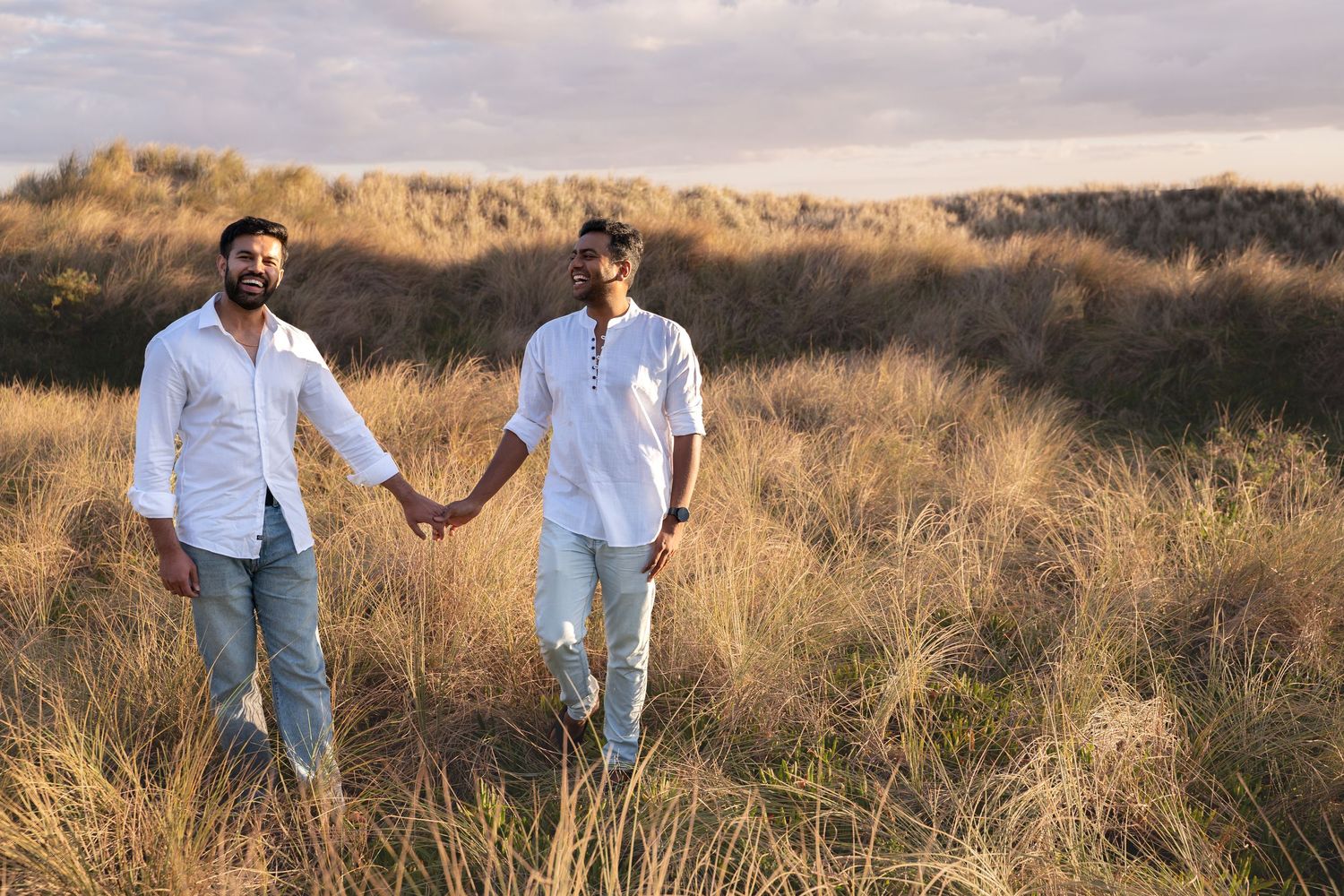 Two men holding hands walking through golden grass fields wearing white shirts and jeans at sunset.