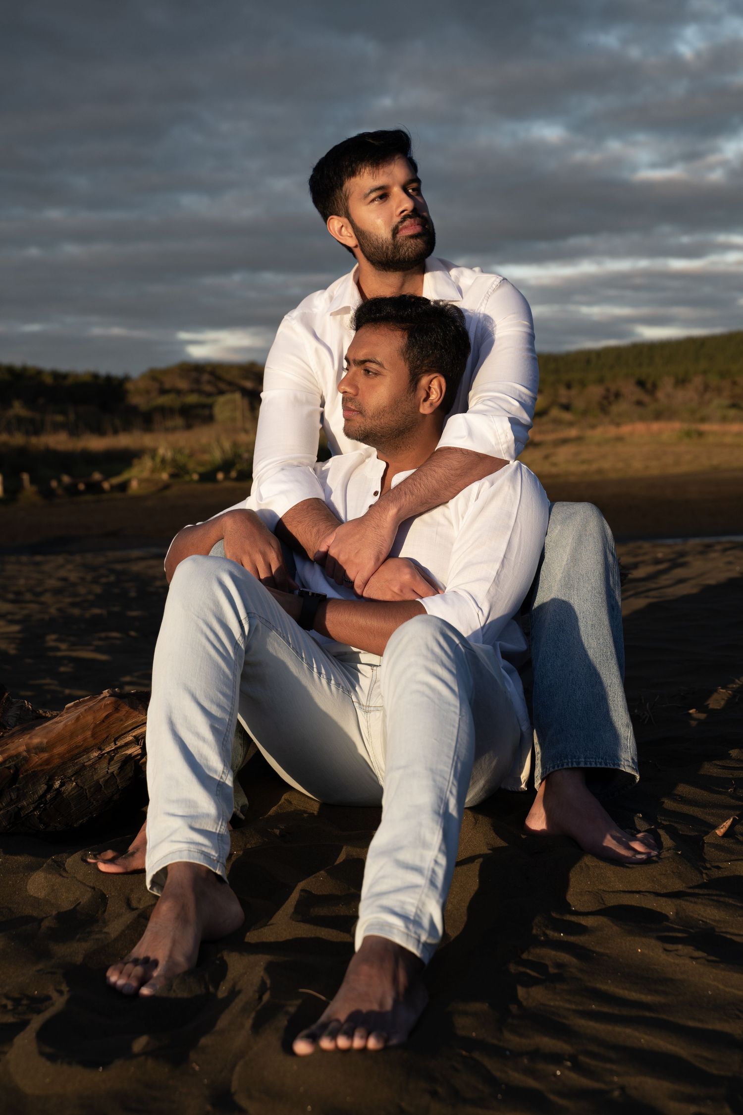 Two men in white shirts and jeans sit together on driftwood at a beach during golden hour.