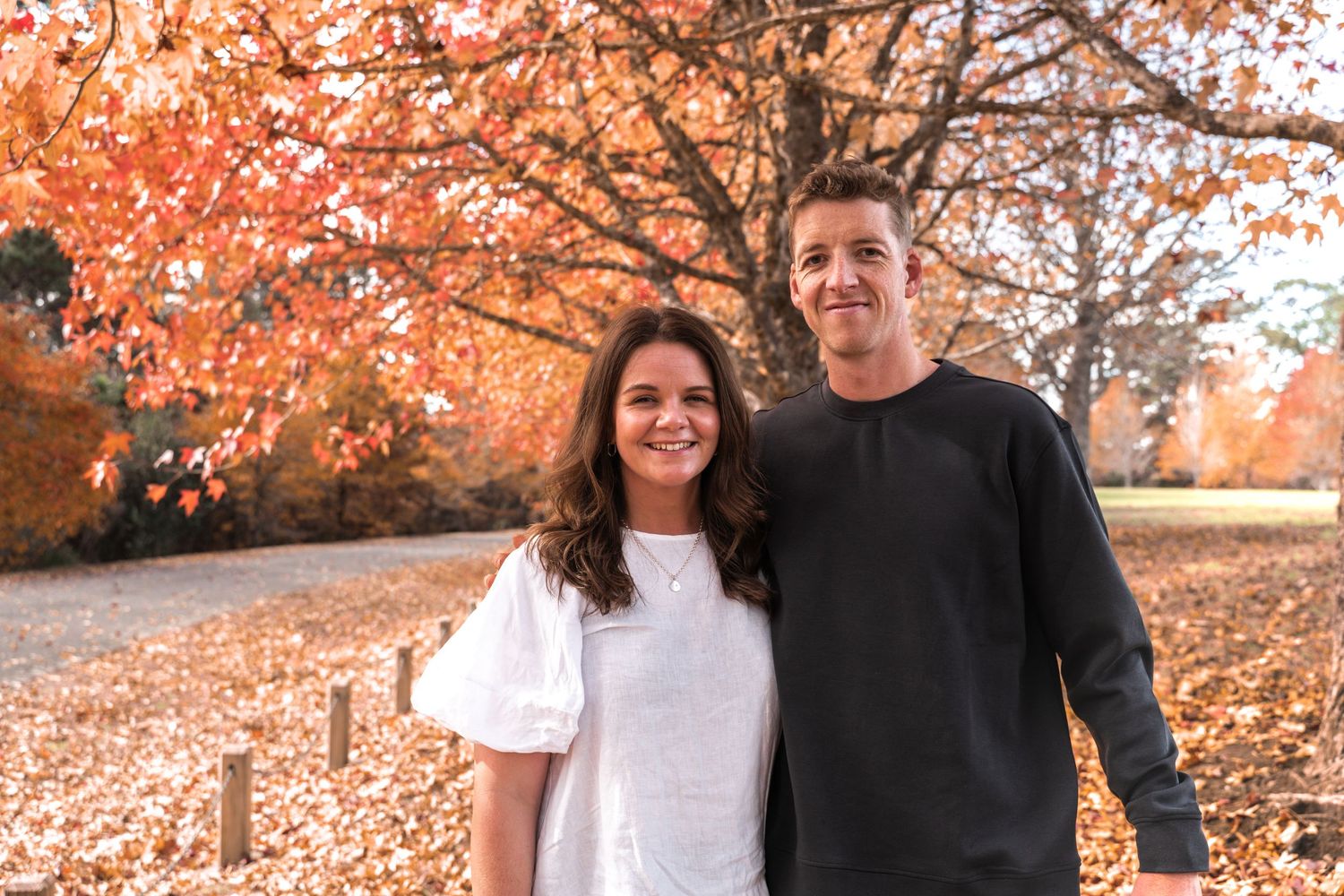 Couple smiling together outdoors in autumn park with vibrant orange and red foliage in background.