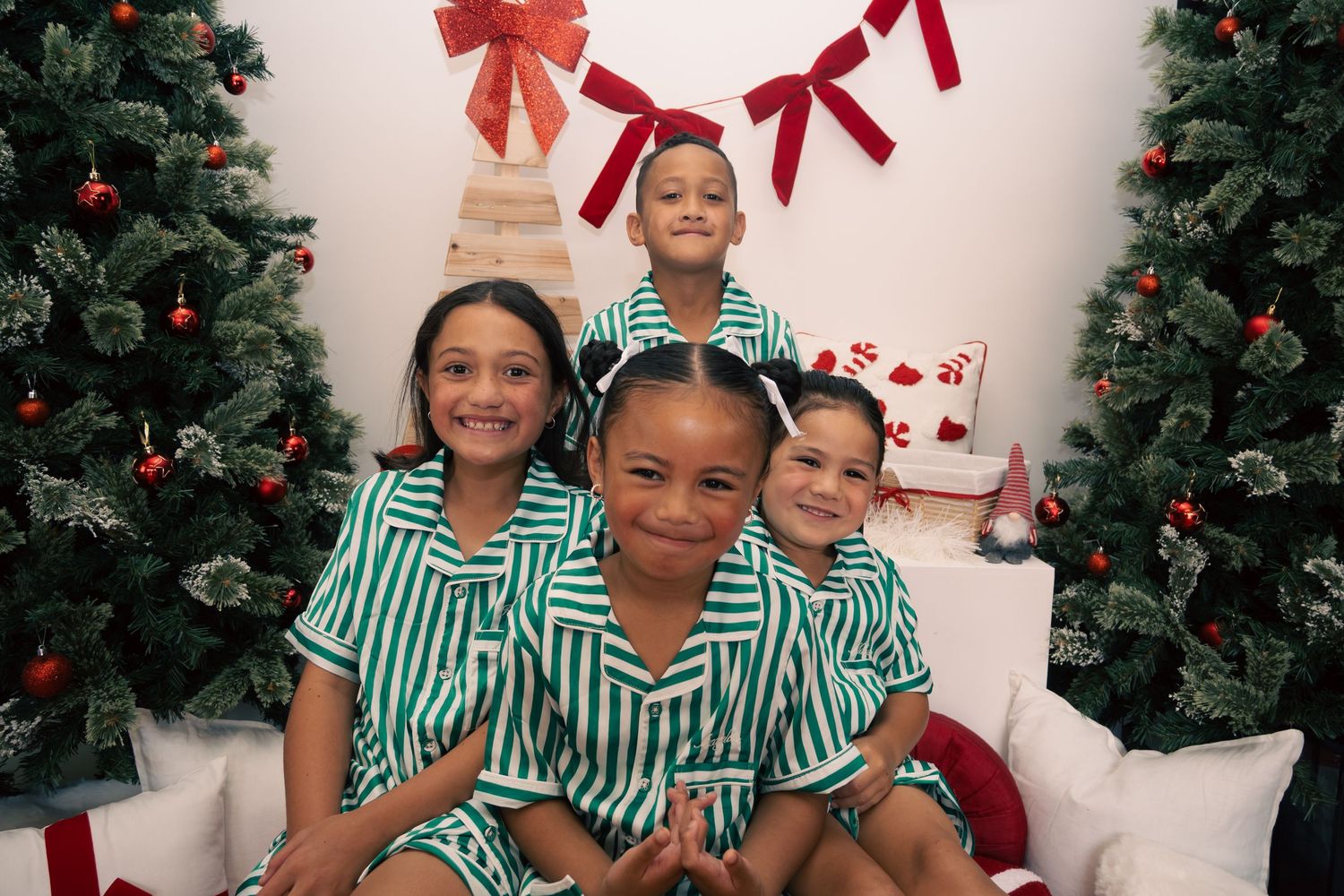 Four children in matching green striped pajamas pose together between Christmas trees with red bows and ornaments.