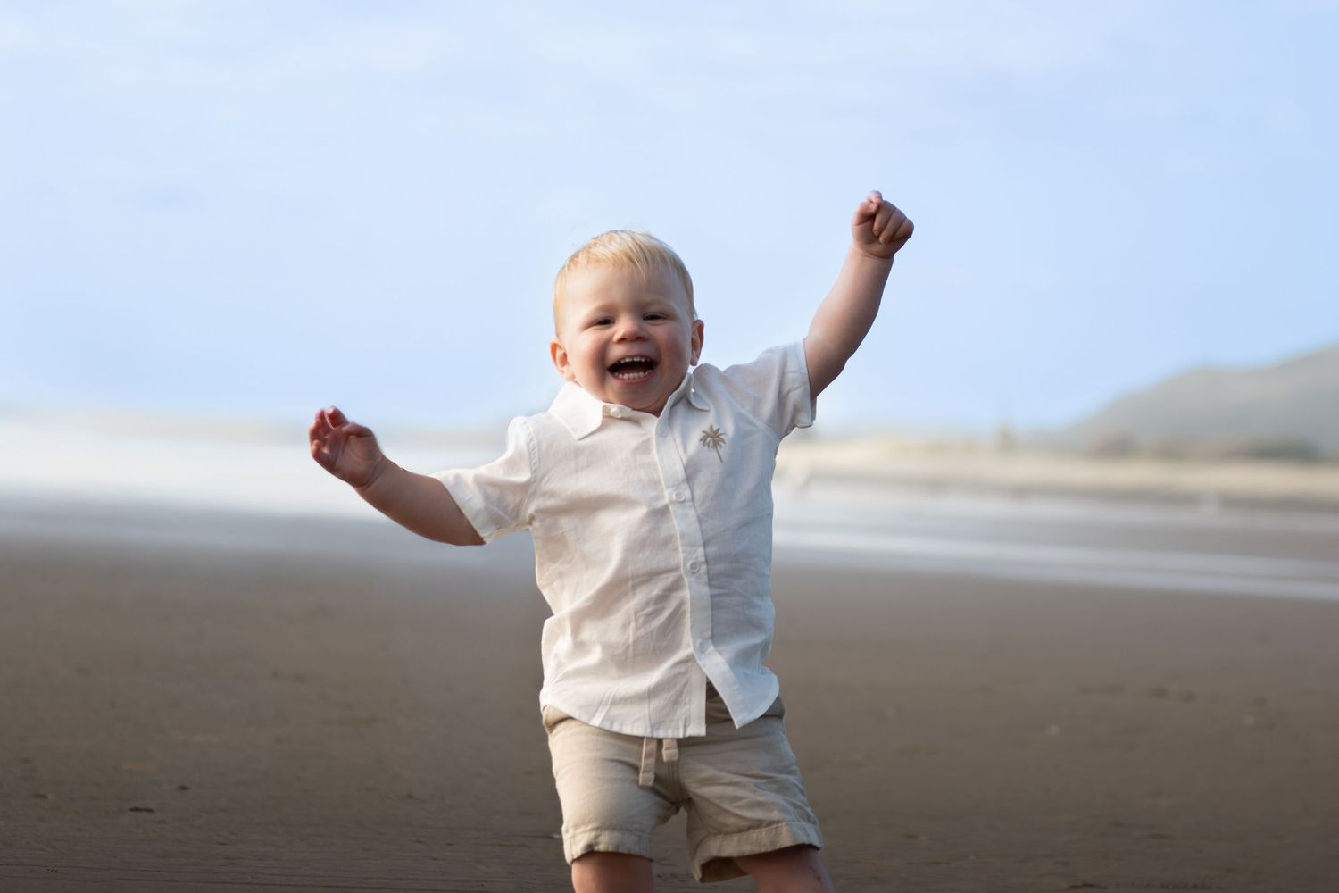 Happy toddler boy in white shirt and khaki shorts celebrating on a sandy beach with arms raised in joy.
