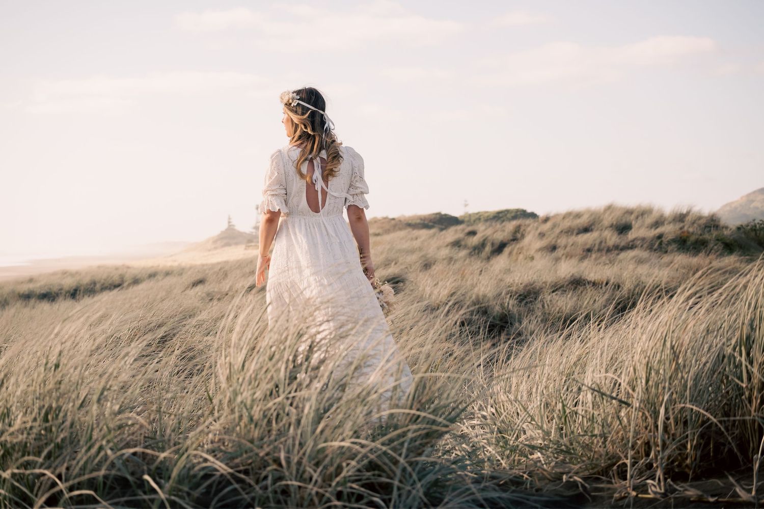 Woman in white boho dress with floral crown walks through golden beach grass dunes at sunset.