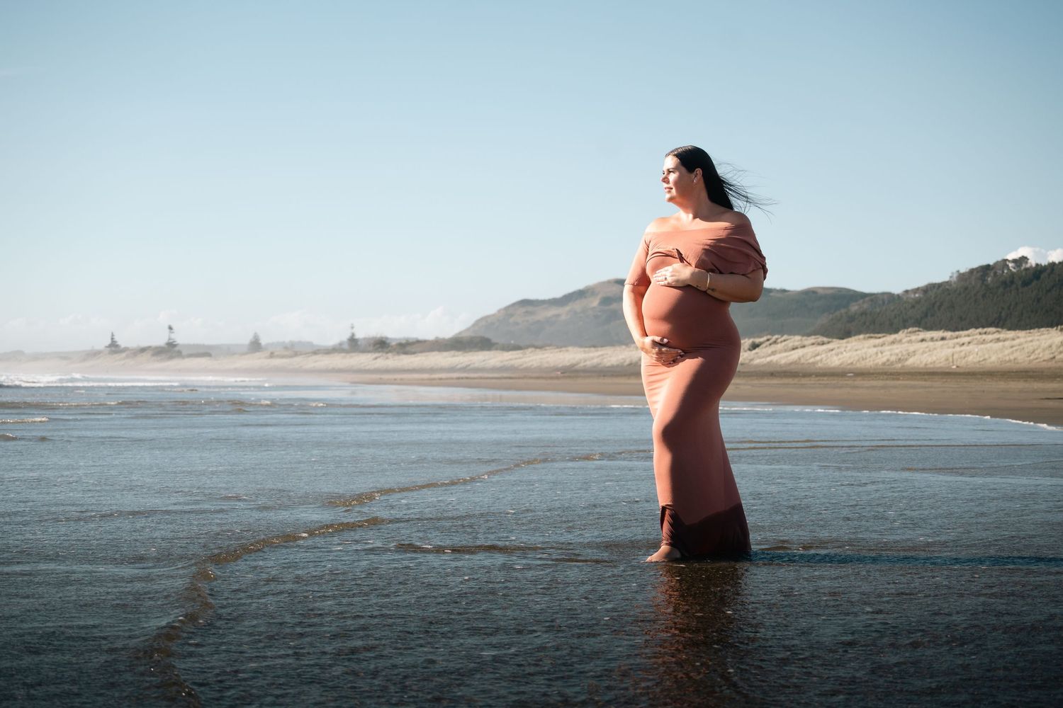 Pregnant woman in pink dress standing in ocean waves on a sandy beach with hills in the background.