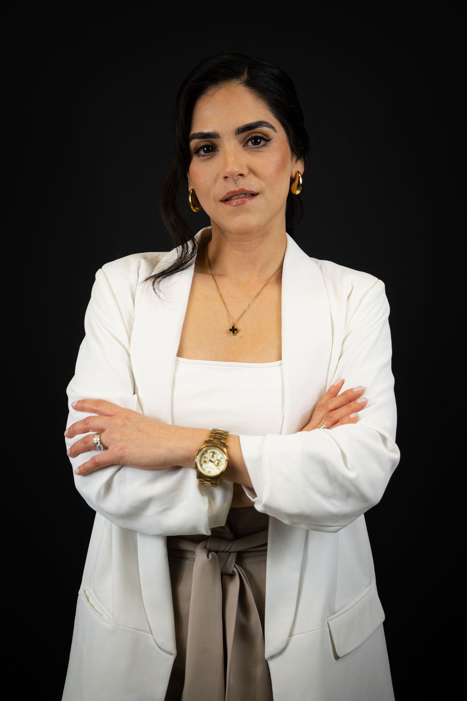 Professional woman in white blazer with arms crossed, wearing gold watch and necklace, against dark background.