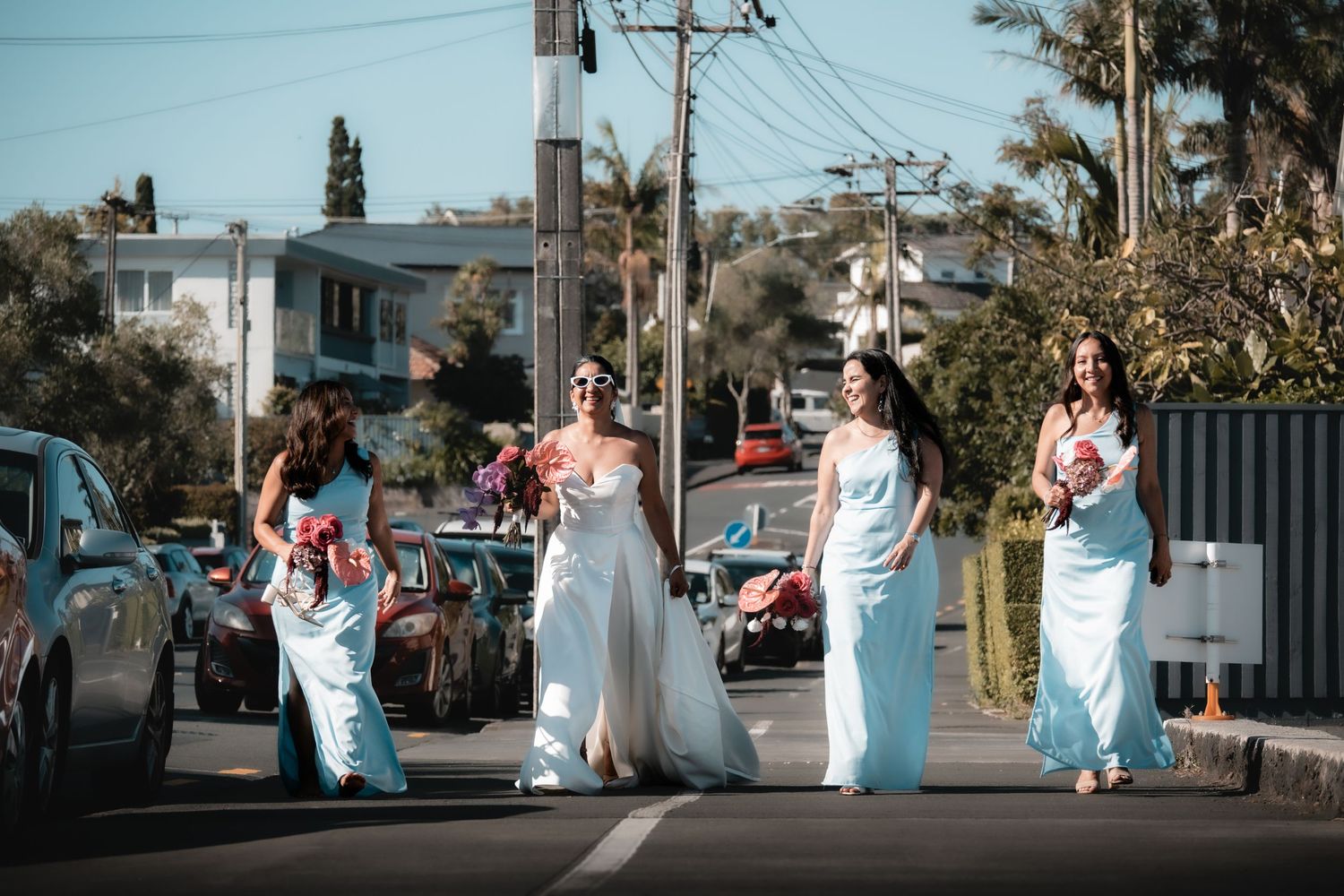 Bride and bridesmaids in light blue gowns walking down a sunny street holding bouquets on a wedding day.