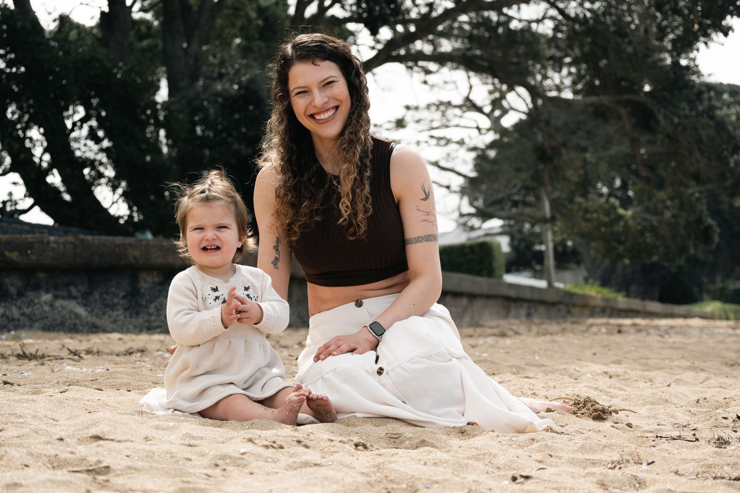 A smiling woman and toddler girl sit together on a sandy beach, with lush green trees in the background.