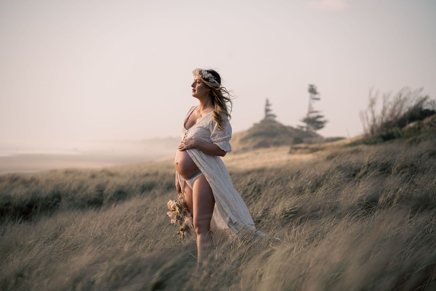Pregnant woman in flowing white dress holding flowers stands in golden grass field at sunset.