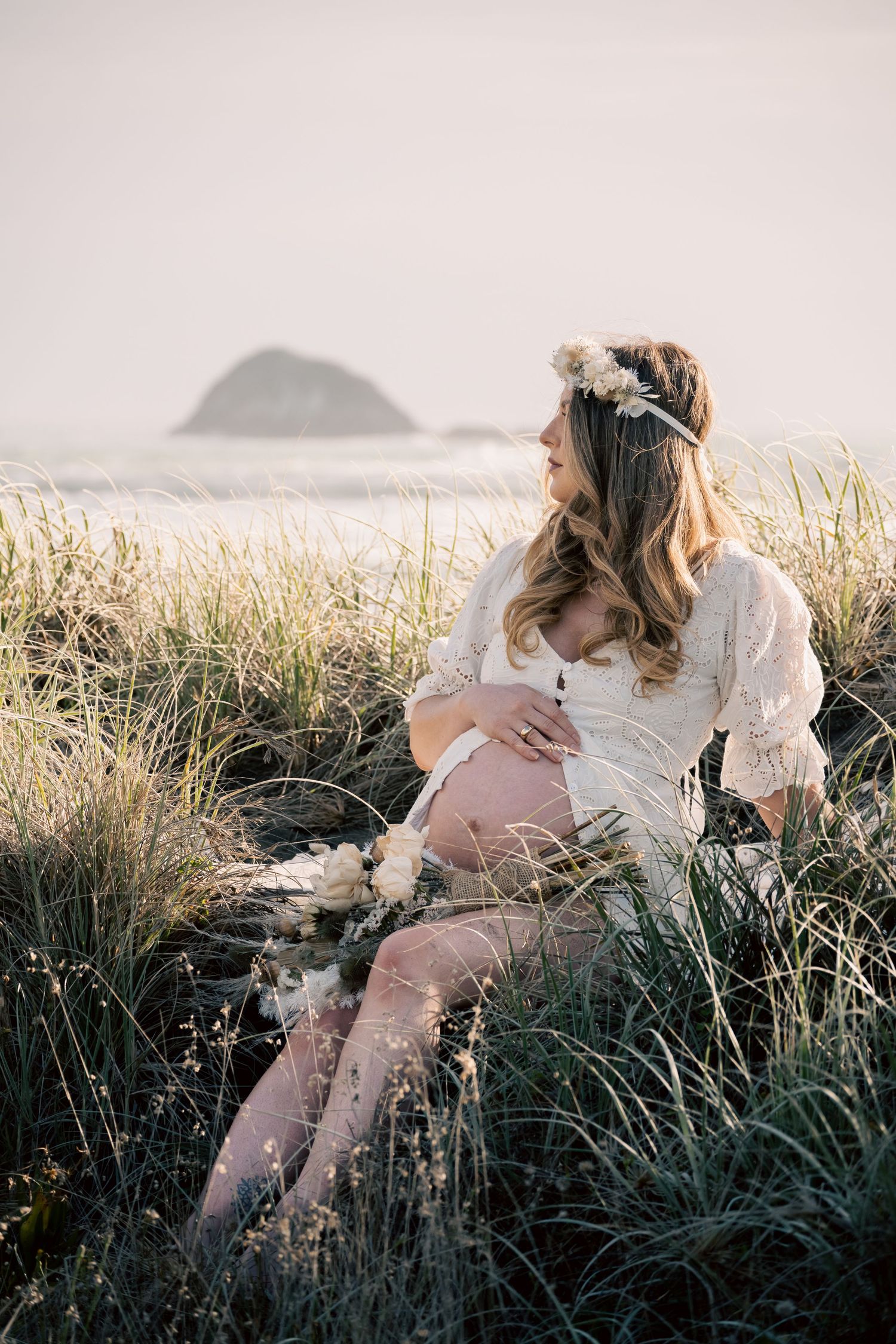 Pregnant woman in white lace dress and floral crown sitting in beach grass, cradling baby bump with ocean view.