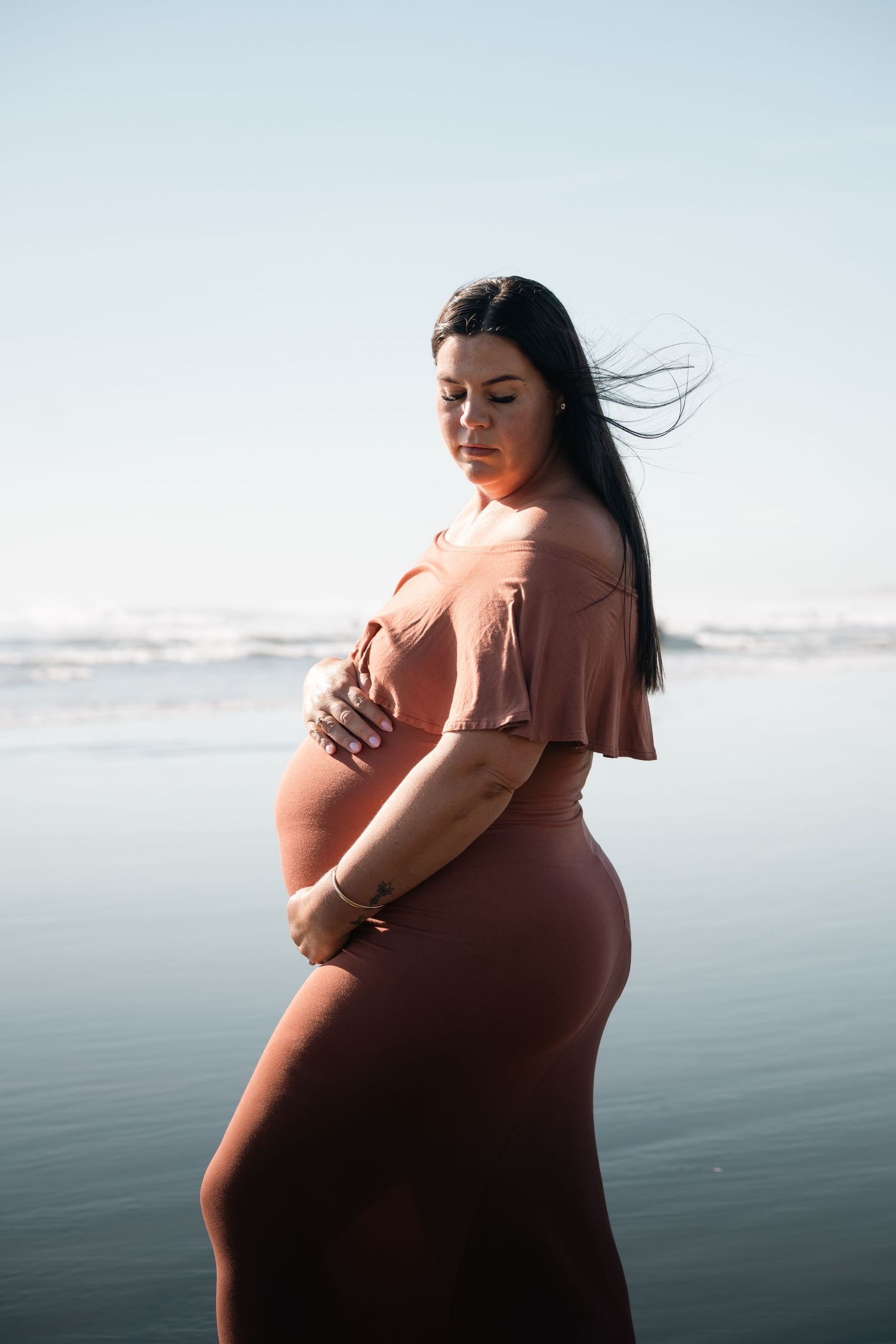 Pregnant woman in rust dress cradles baby bump on beach, hair blowing in wind, ocean backdrop.