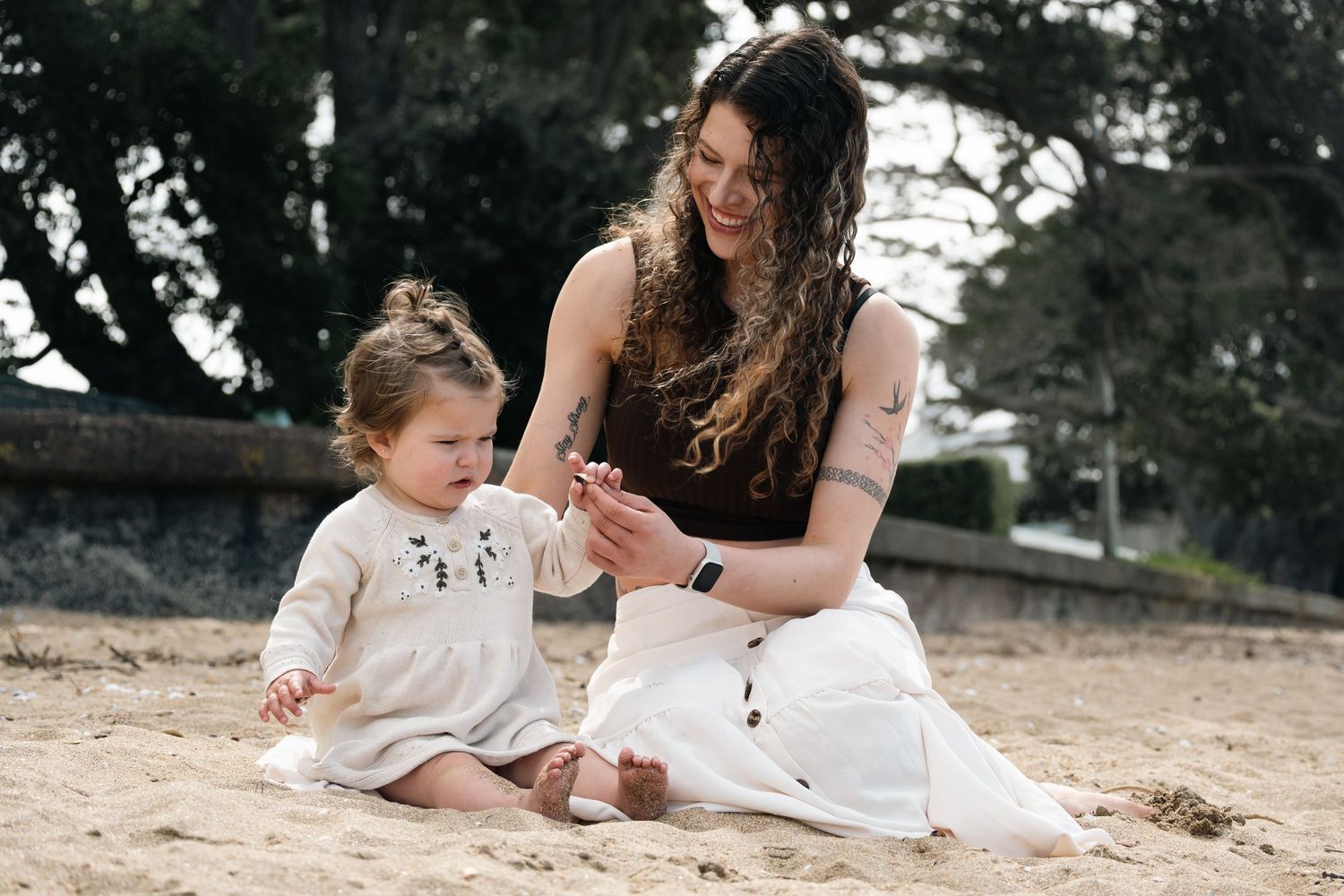 A young mother and toddler girl sit together on sandy beach, sharing a tender moment outdoors.