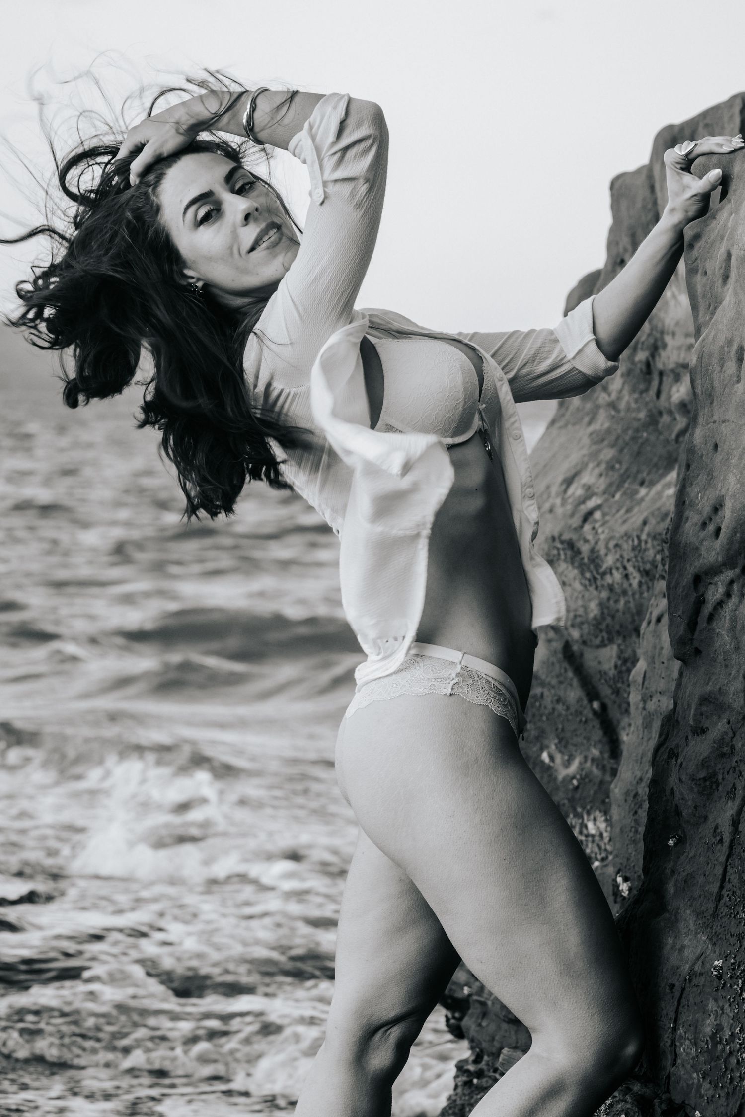 Black and white photo of a woman in lingerie posing against rocky seaside cliffs with wind-blown hair.