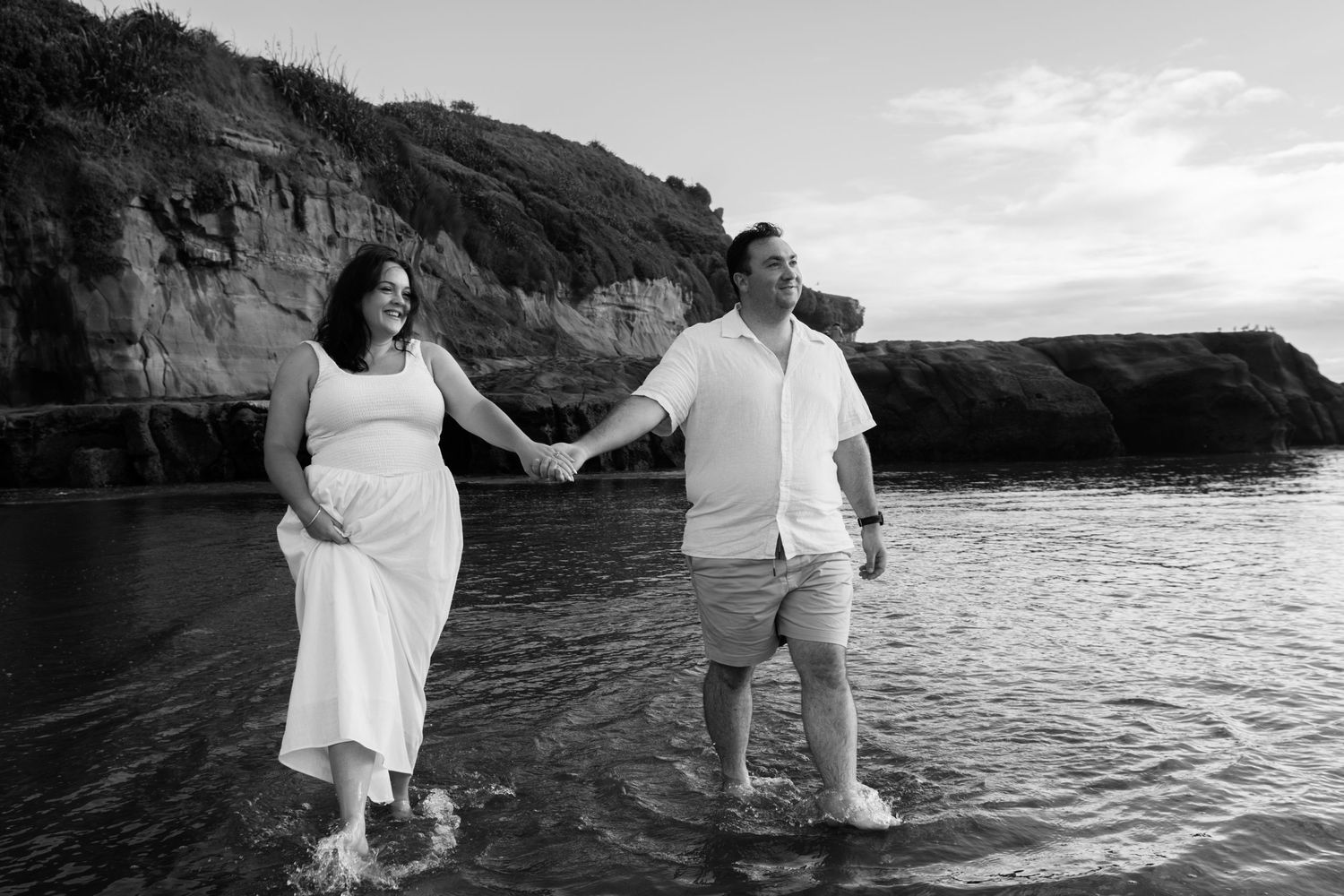 Pregnant couple holds hands walking in ocean waves near rocky cliffs in black and white beach maternity photo.