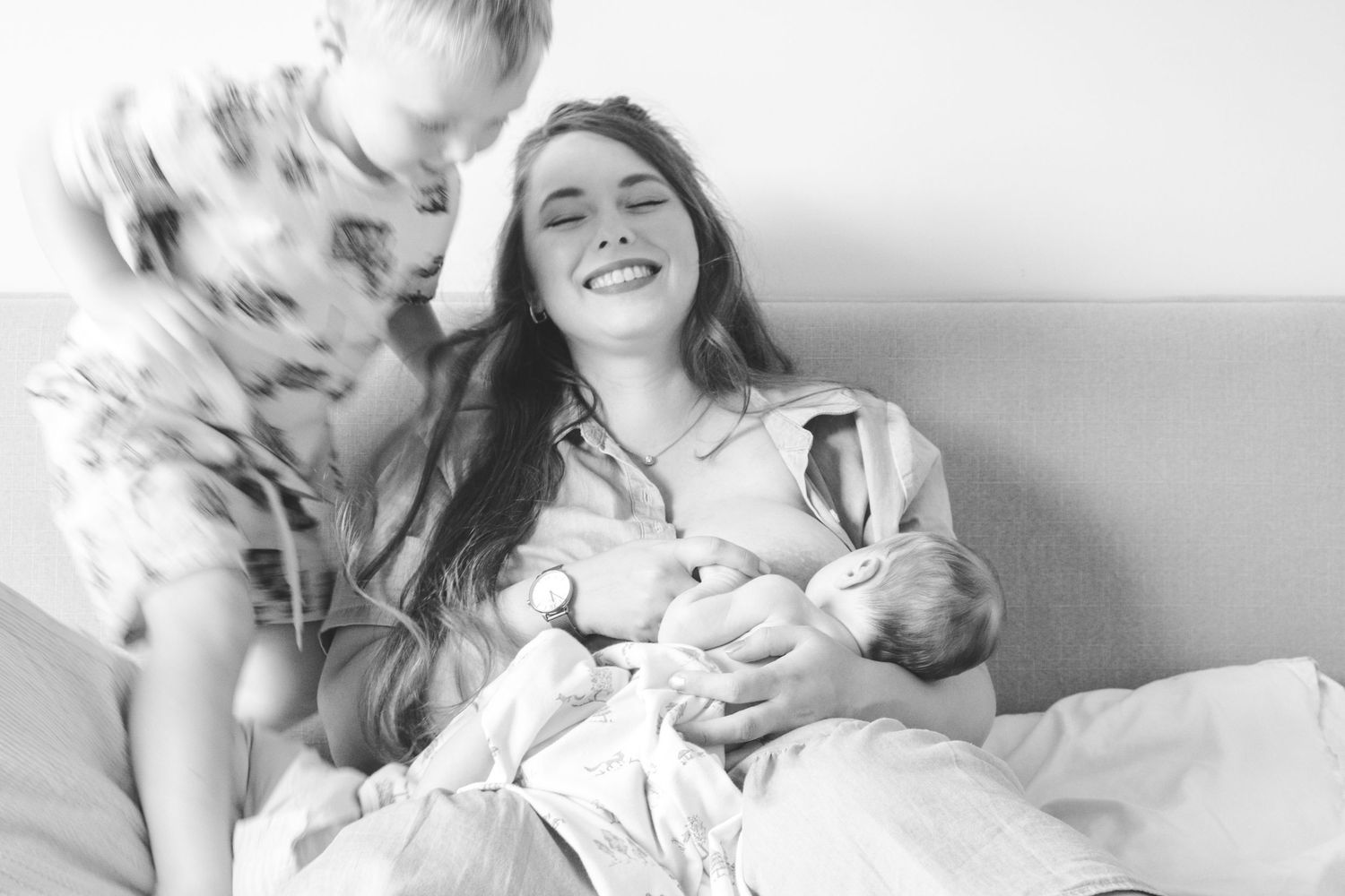 Black and white photo of a smiling mother breastfeeding newborn baby in hospital bed with loved one nearby.