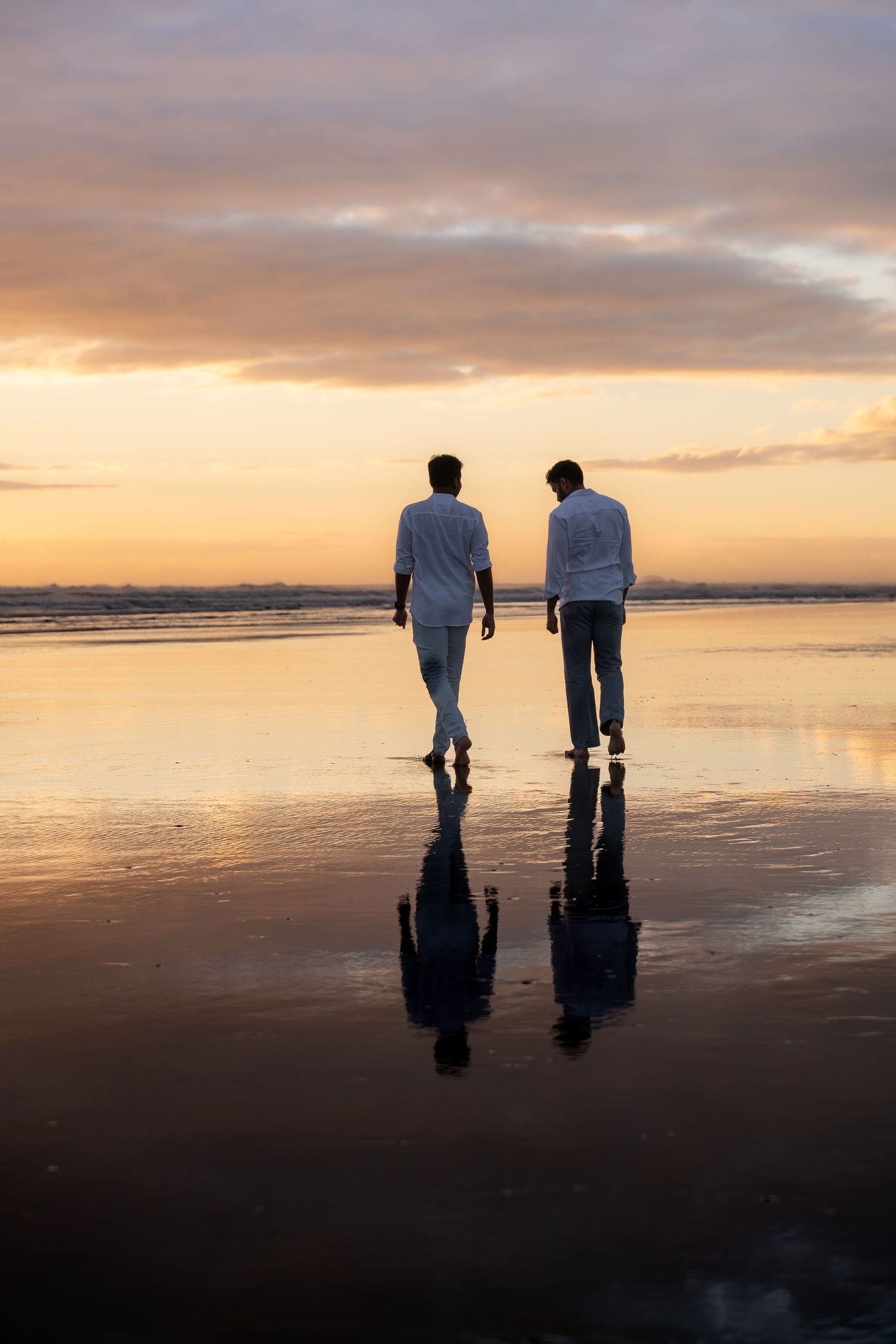 Two men walking on a reflective beach at sunset, wearing white shirts, with golden sky and water reflections.