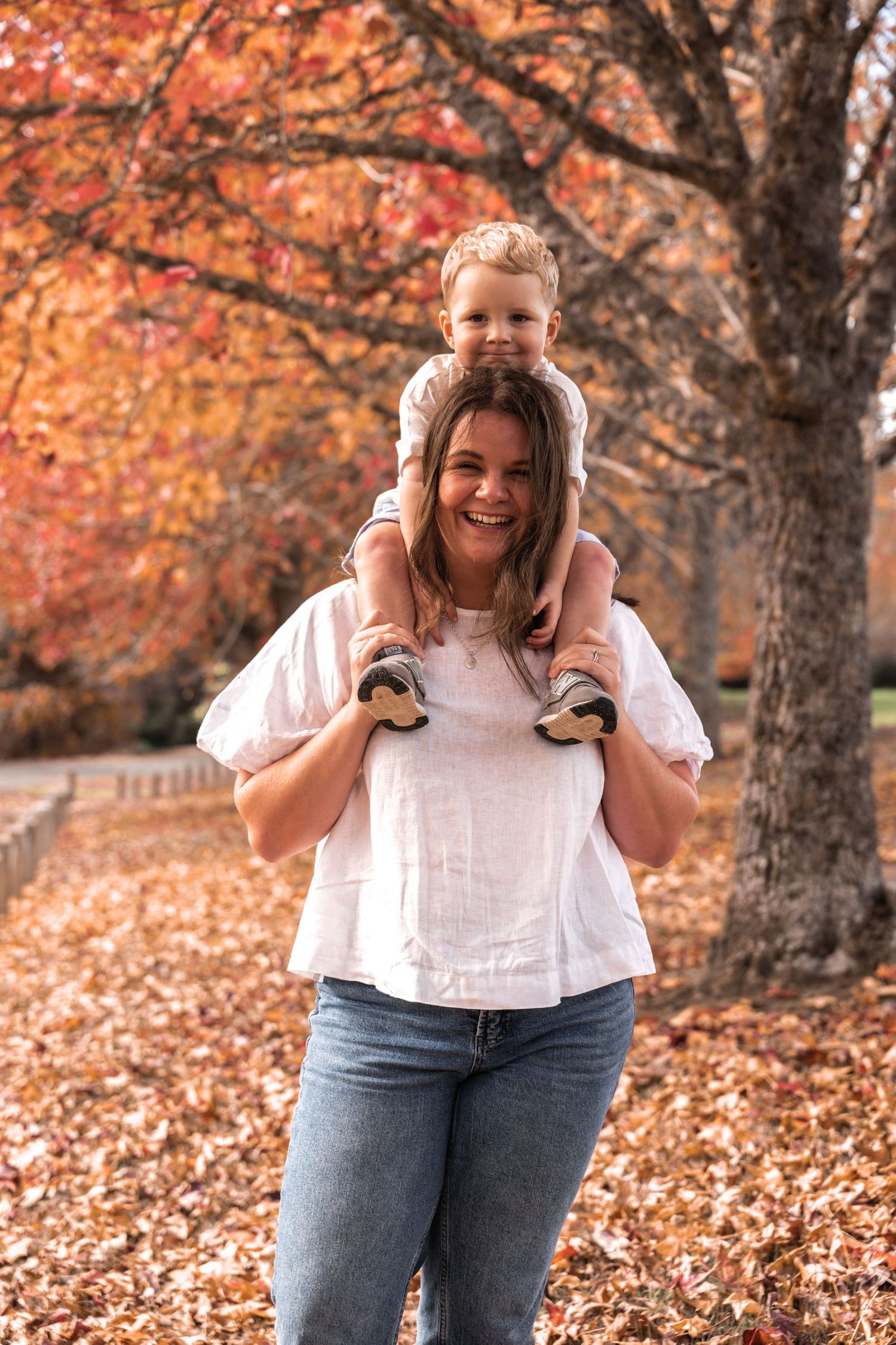 A smiling woman carries a young child on her shoulders among autumn trees with colorful fallen leaves.