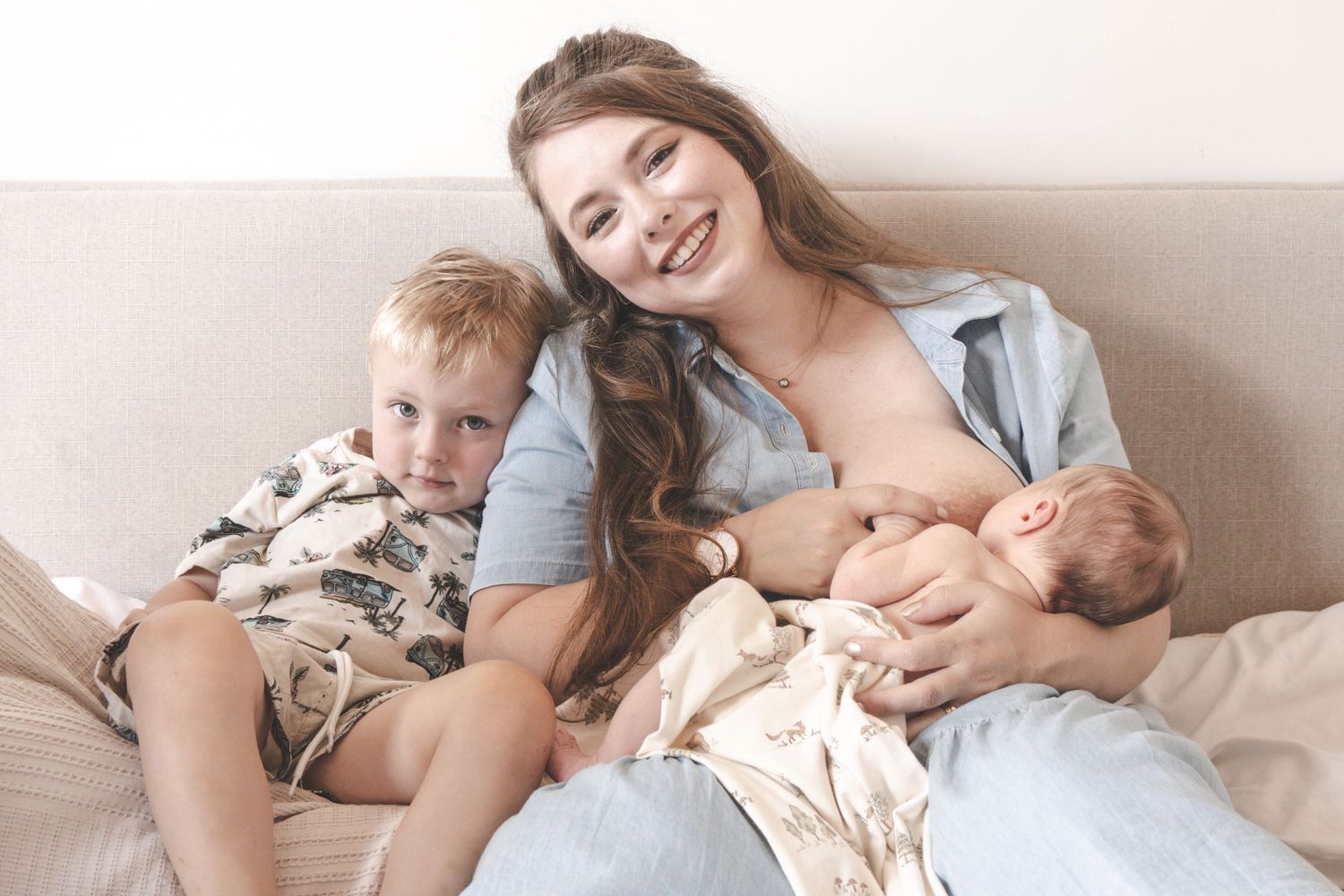 Smiling mother breastfeeding newborn baby while sitting on bed with young toddler son beside her.