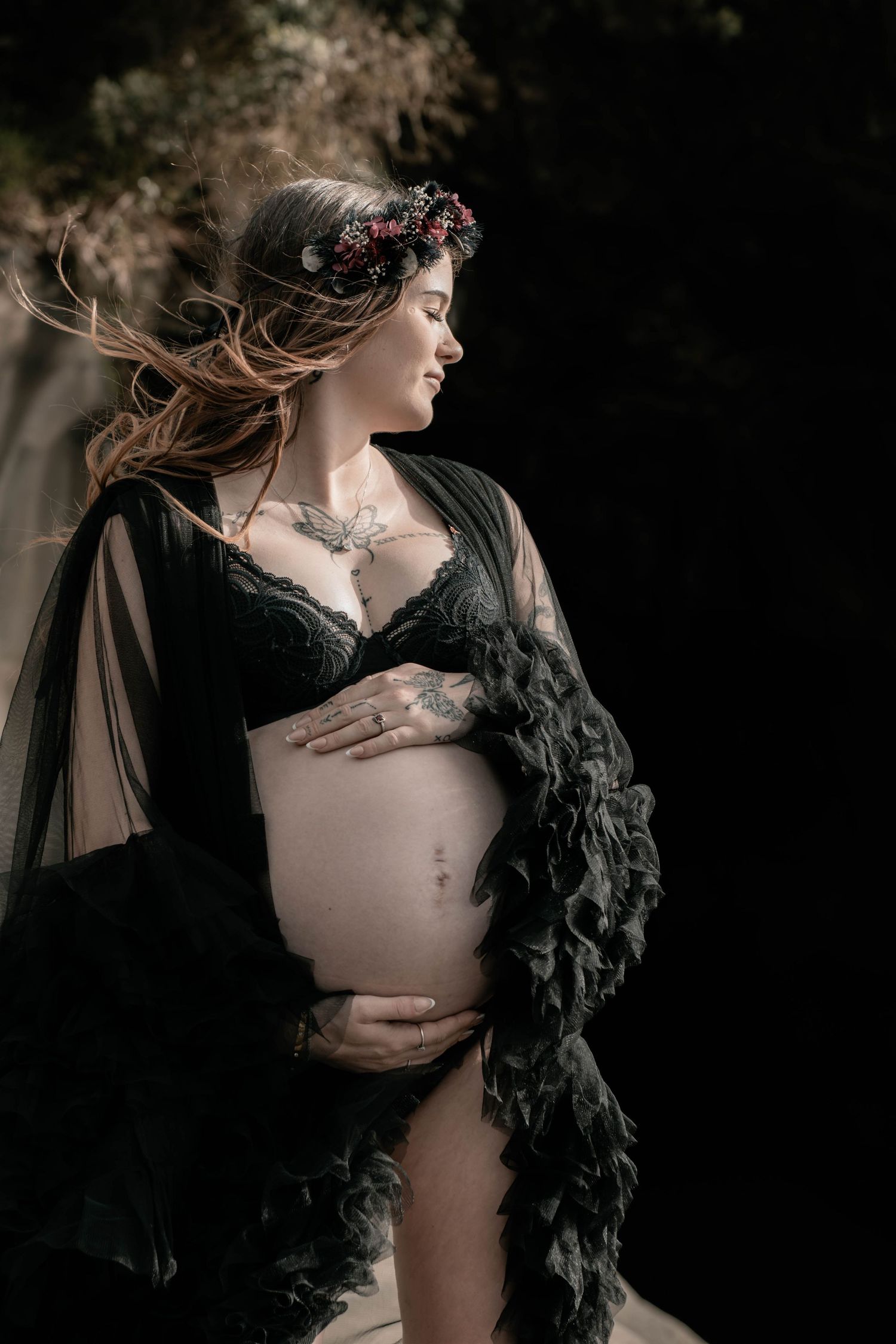 Pregnant woman in black lace bra and sheer robe with floral crown, cradling baby bump outdoors.