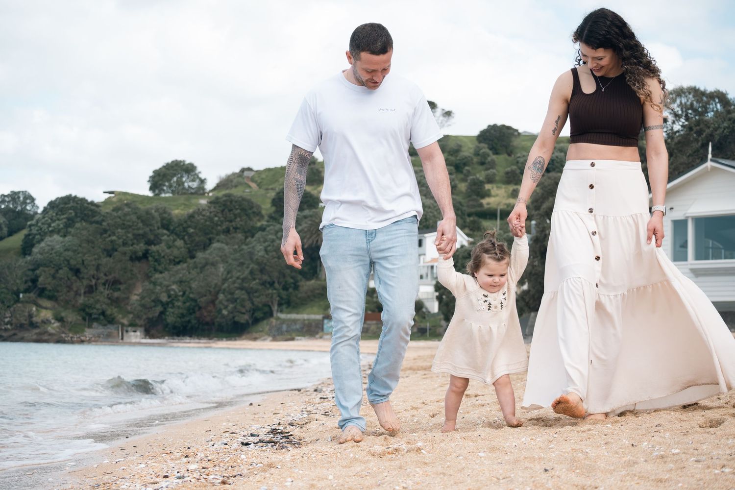 A happy family of three walks along a sandy beach, parents holding their toddler's hands near calm ocean waters.
