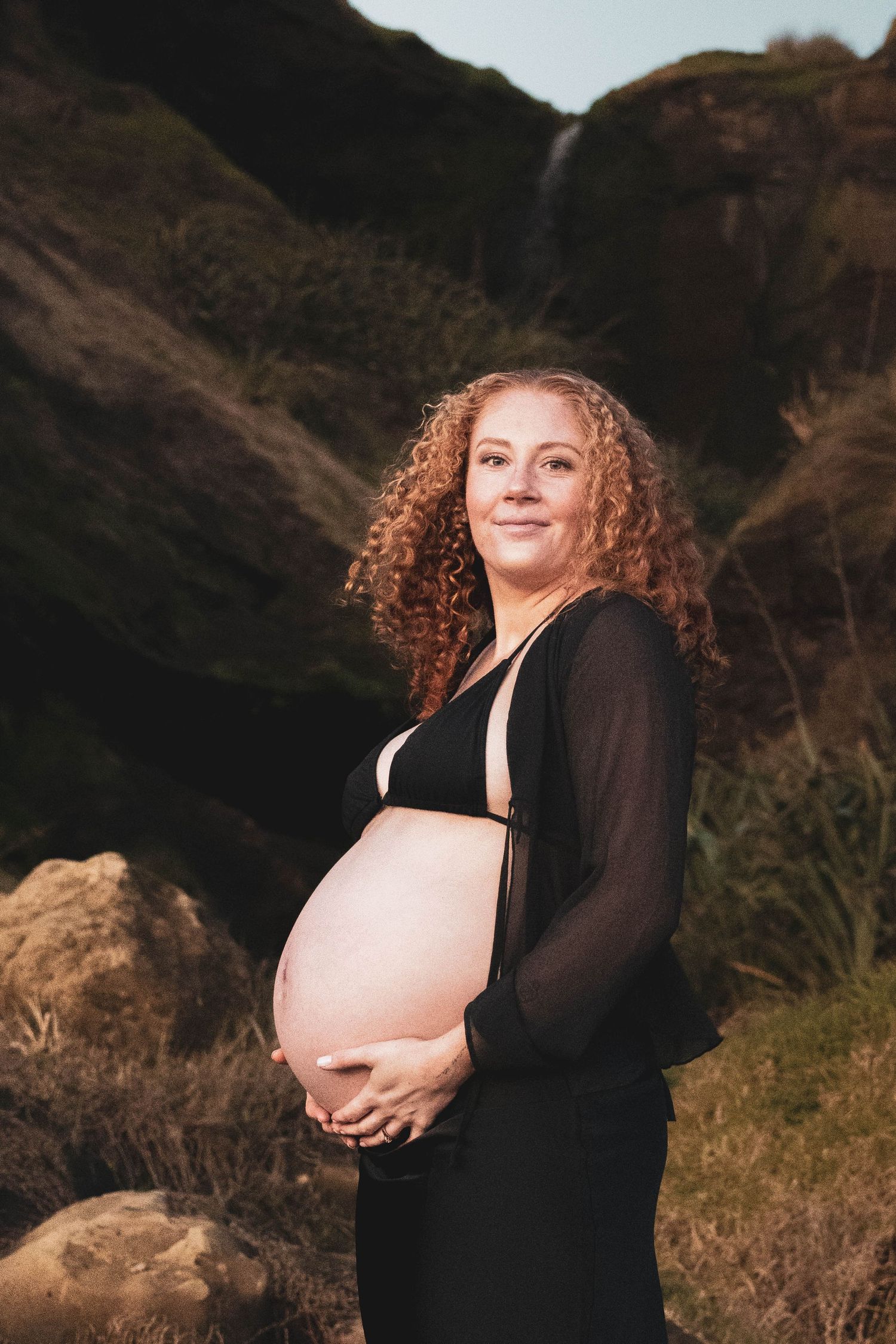 Pregnant woman with curly red hair holding her bare baby bump outdoors near a rocky waterfall backdrop.