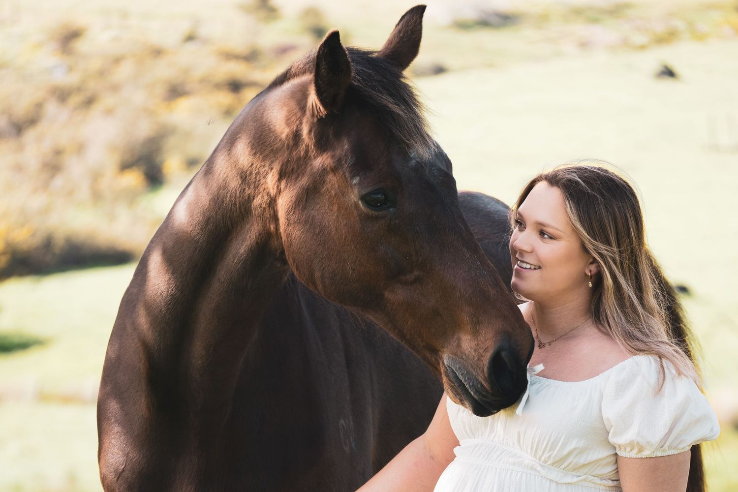 A smiling woman in a white dress stands beside a dark brown horse in a sunny green field.