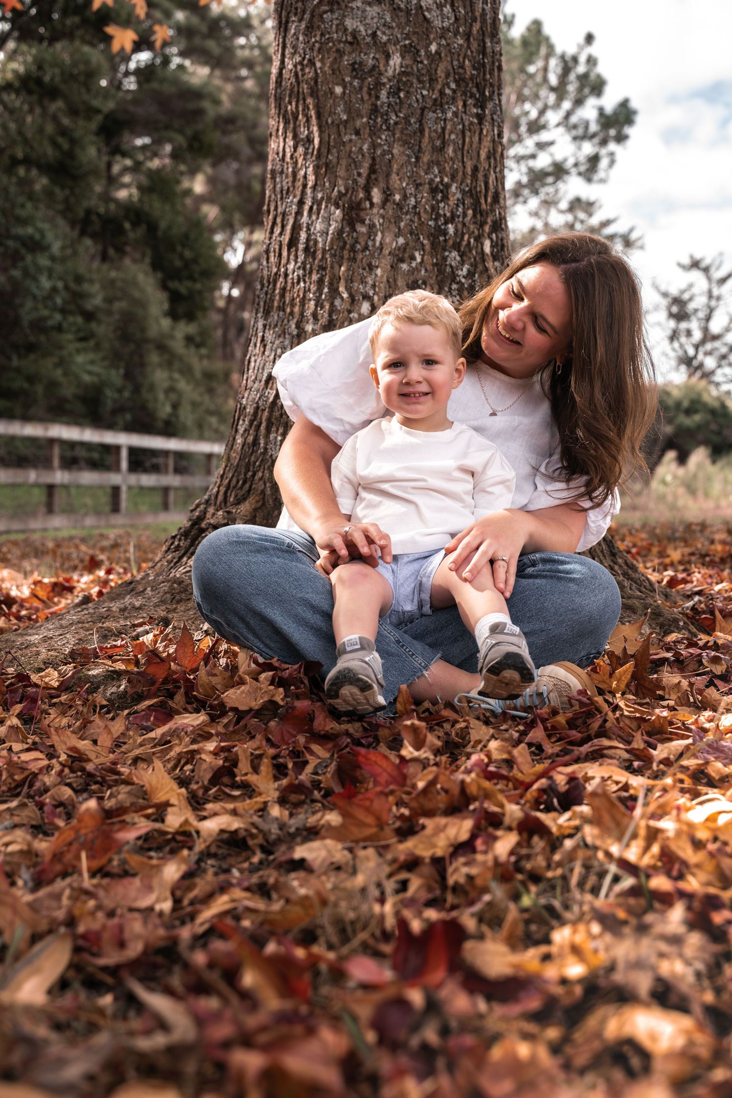 A mother and toddler son sit together against a tree trunk surrounded by autumn leaves, smiling joyfully outdoors.