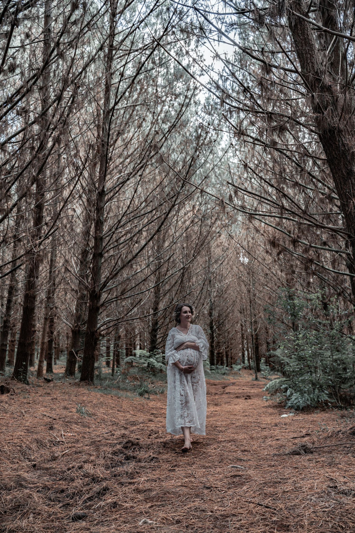 Pregnant woman in white lace dress cradling baby bump while standing on pine needle path in moody forest.