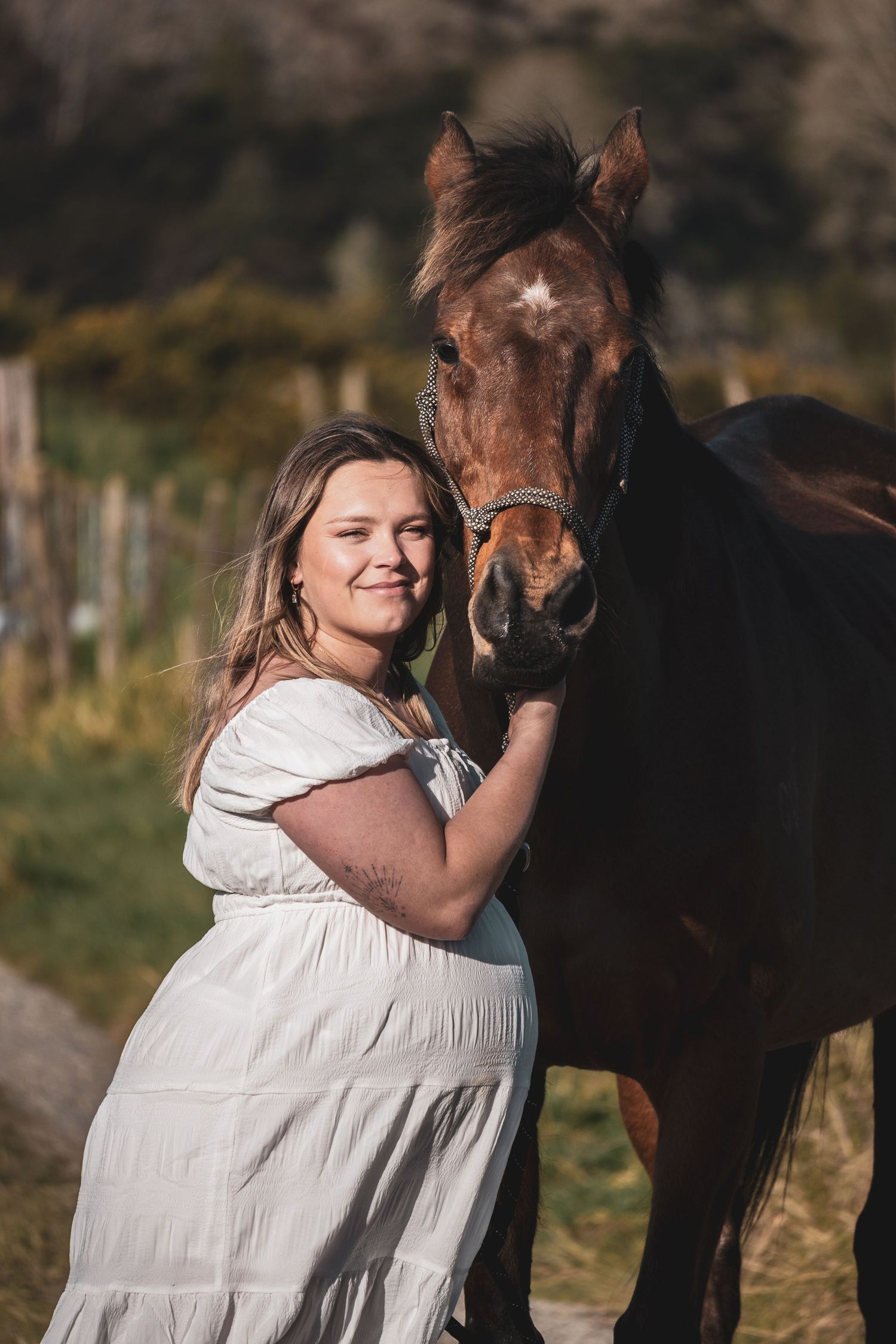 Woman in white dress smiling beside a brown horse outdoors in a sunny field.