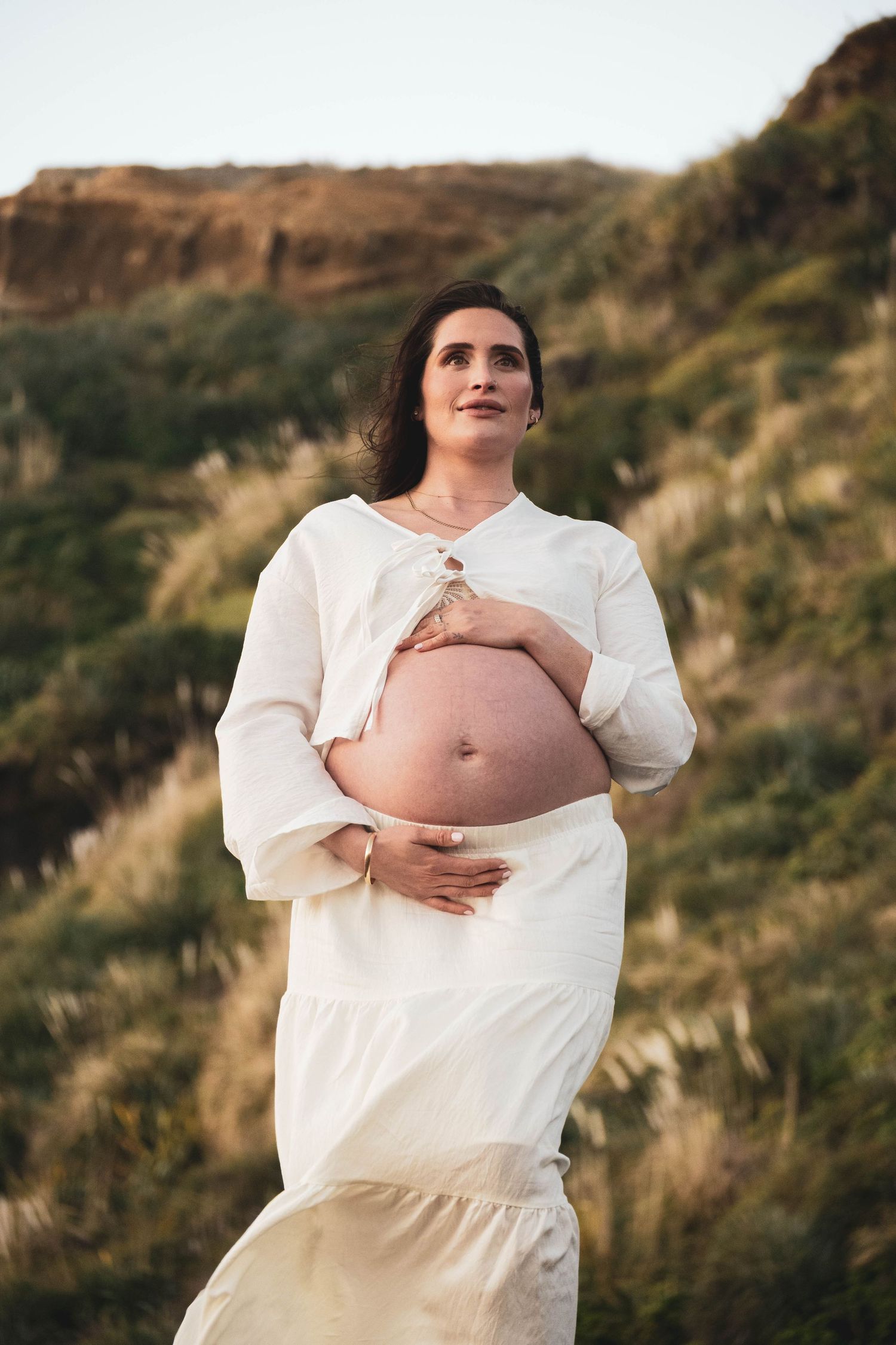 Pregnant woman in white dress cradling bare baby bump outdoors with green hillside backdrop.