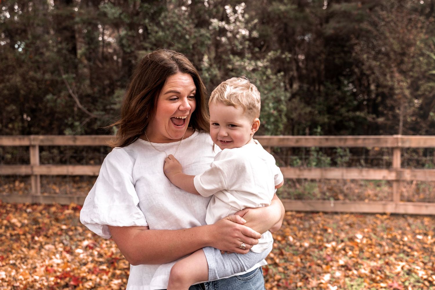 A woman in a white top laughs while holding a smiling toddler outdoors among autumn leaves near a wooden fence.