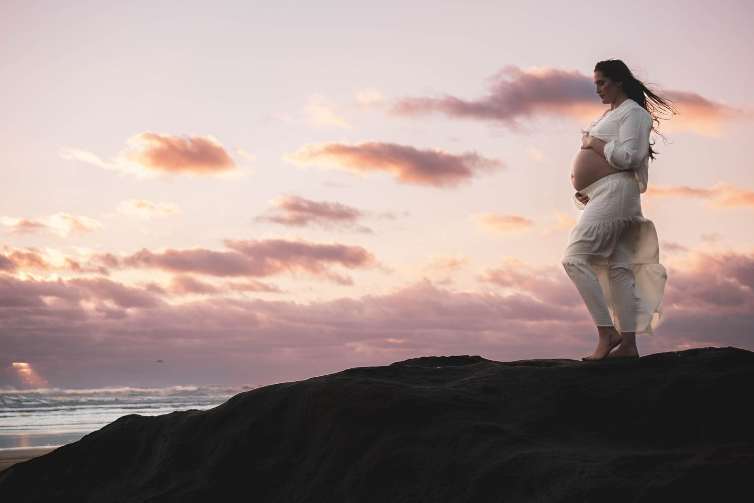 Pregnant woman in white dress standing on rocks at sunset by the ocean during golden hour.