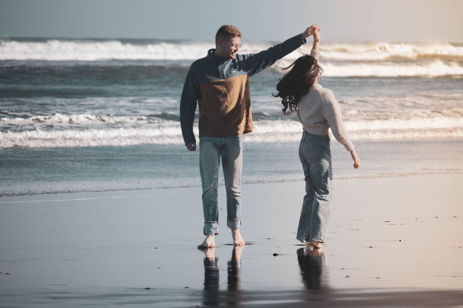Couple dancing barefoot on a wet beach, holding hands with ocean waves in the background on a sunny day.