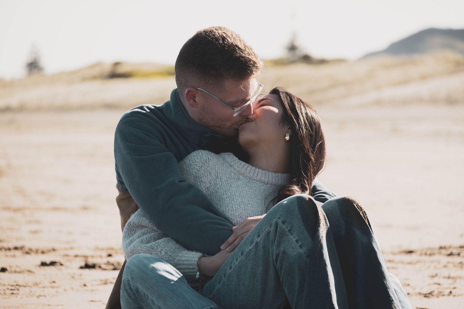 Romantic couple sharing a tender kiss while sitting on a sandy beach on a sunny day.