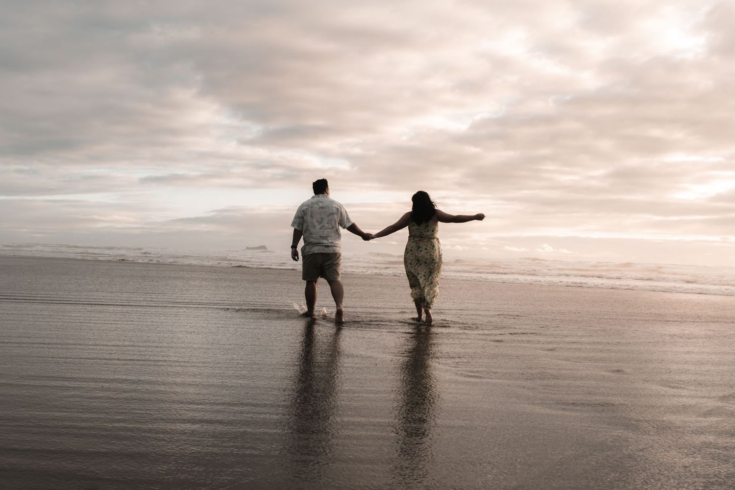 Couple holding hands walking on a scenic beach at sunset with dramatic cloudy skies and wet sand.