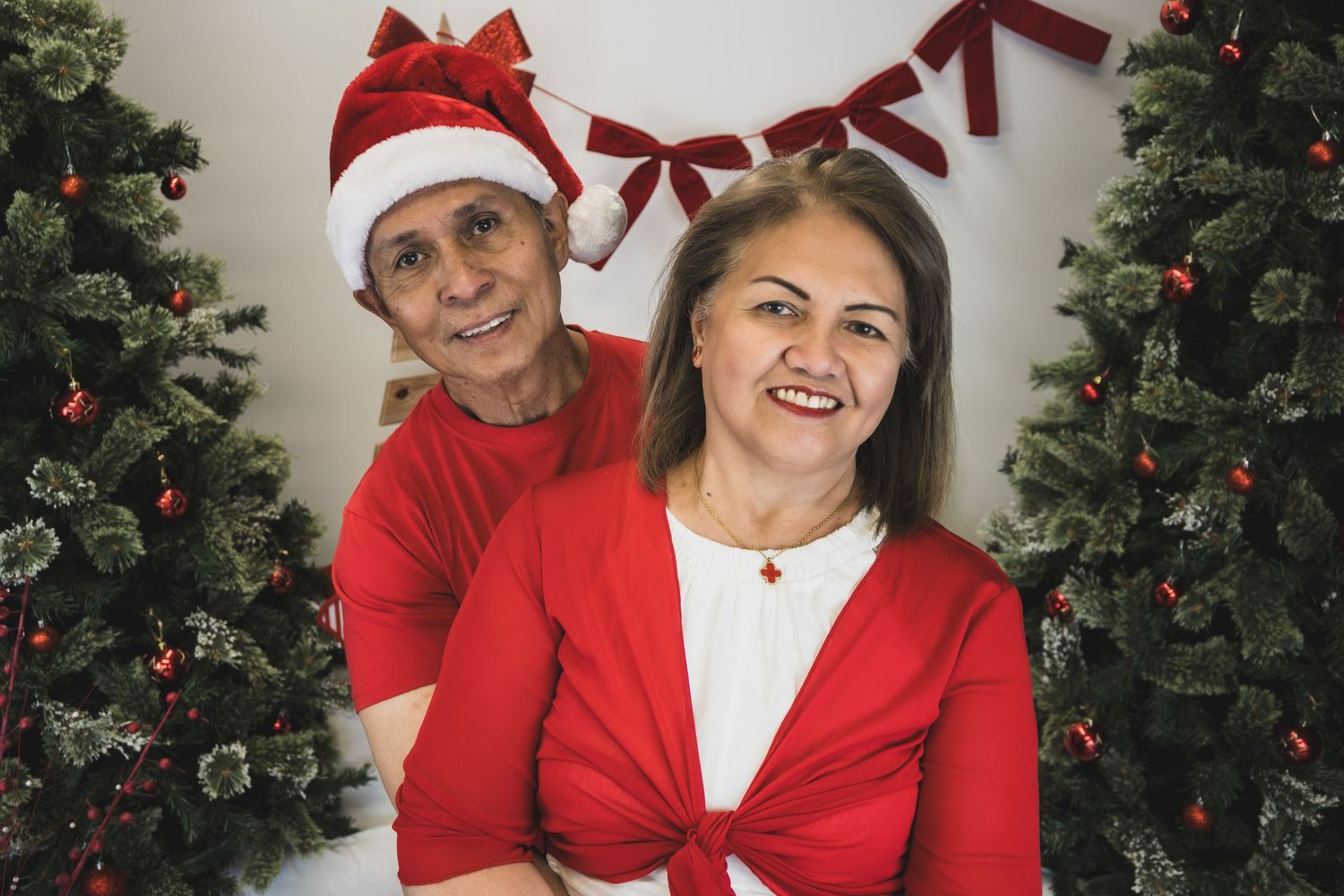 Couple in red Christmas outfits posing between decorated trees, man wearing Santa hat, festive holiday portrait.