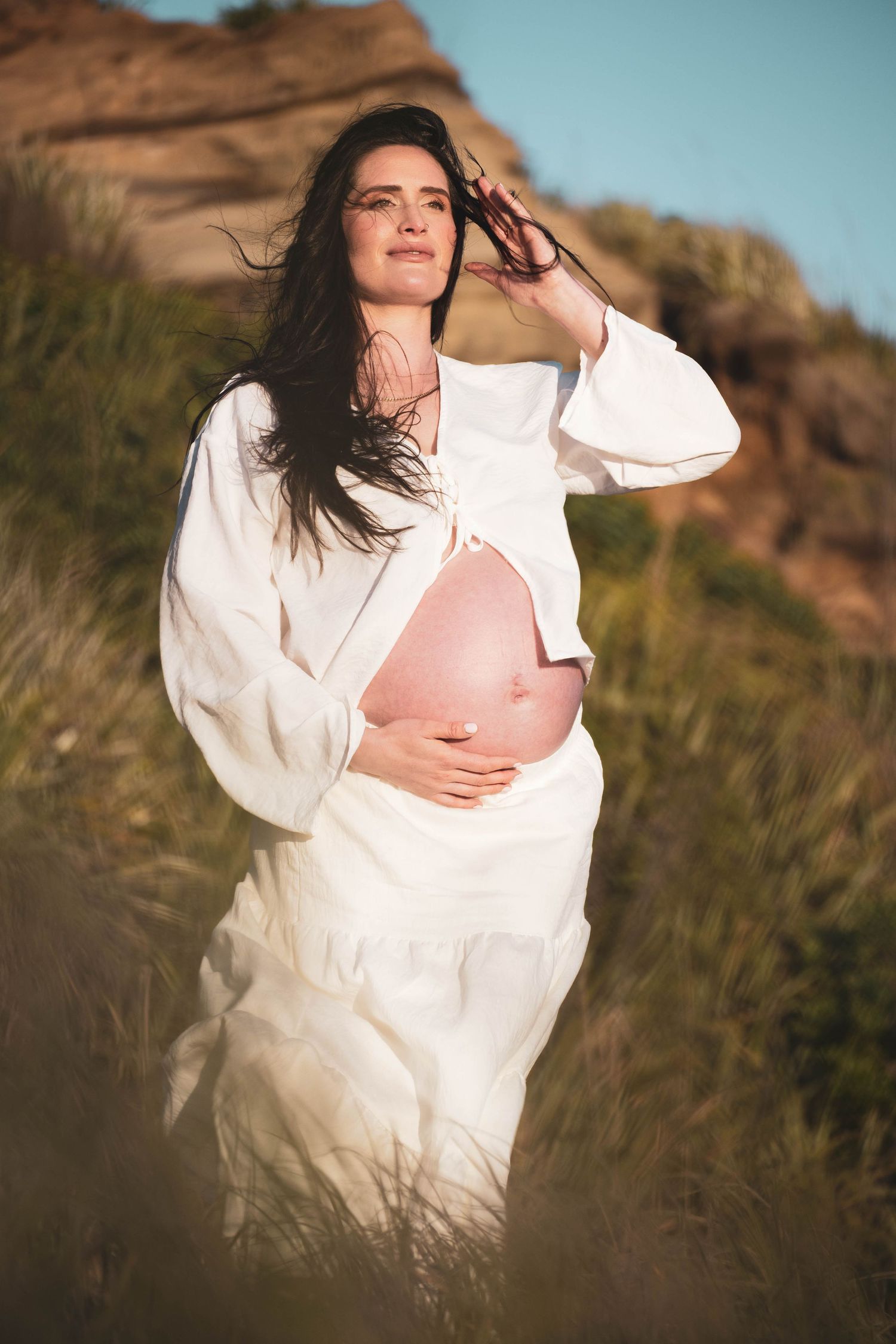 Pregnant woman in white dress outdoors, cradling her bare baby bump amid golden grass and rocky hills in warm sunlight.