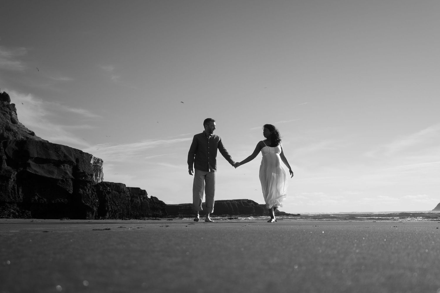 Black and white photo of a couple holding hands on a beach, walking near rocky cliffs under a clear sky.