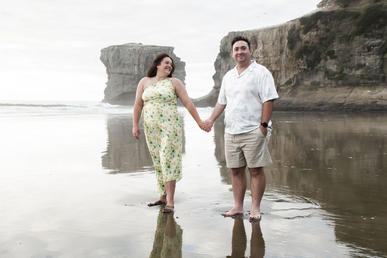Couple holding hands walking barefoot on a reflective beach with dramatic rock formations in the background.