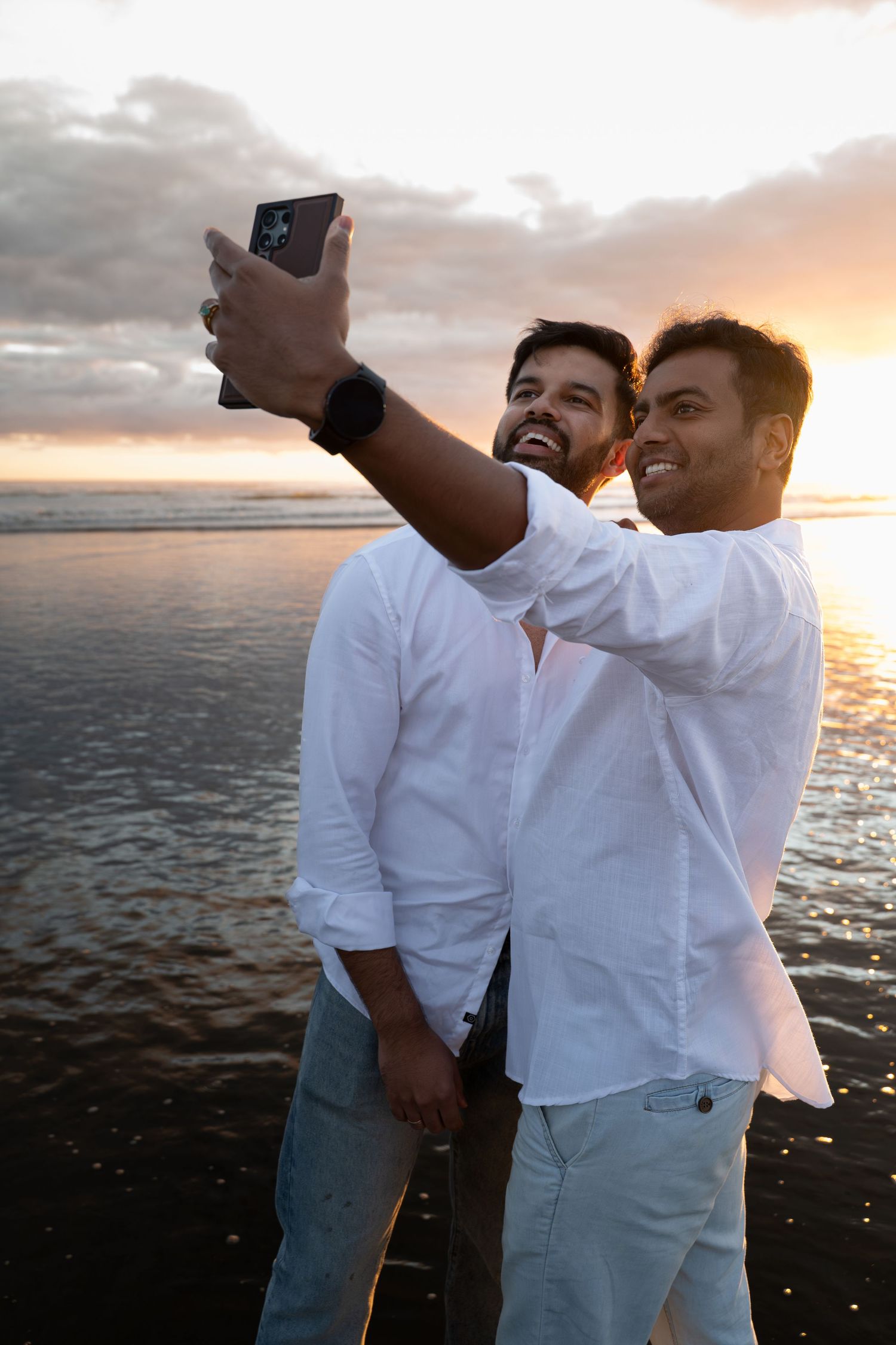 Two men in white shirts take a selfie on a beach at sunset, smiling with ocean waves in the background.